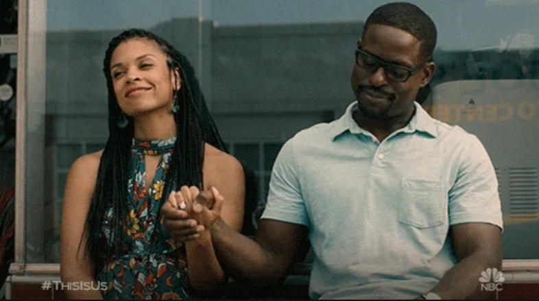 A couple sitting outdoors holds hands and smiles warmly at each other. Both are casually dressed, radiating a sense of contentment and connection