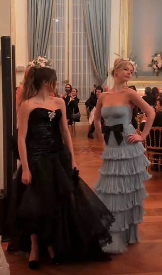 celebrity drama Two people in elegant ball gowns pose indoors; one in a strapless black dress with floral details, and the other in a tiered strapless dress