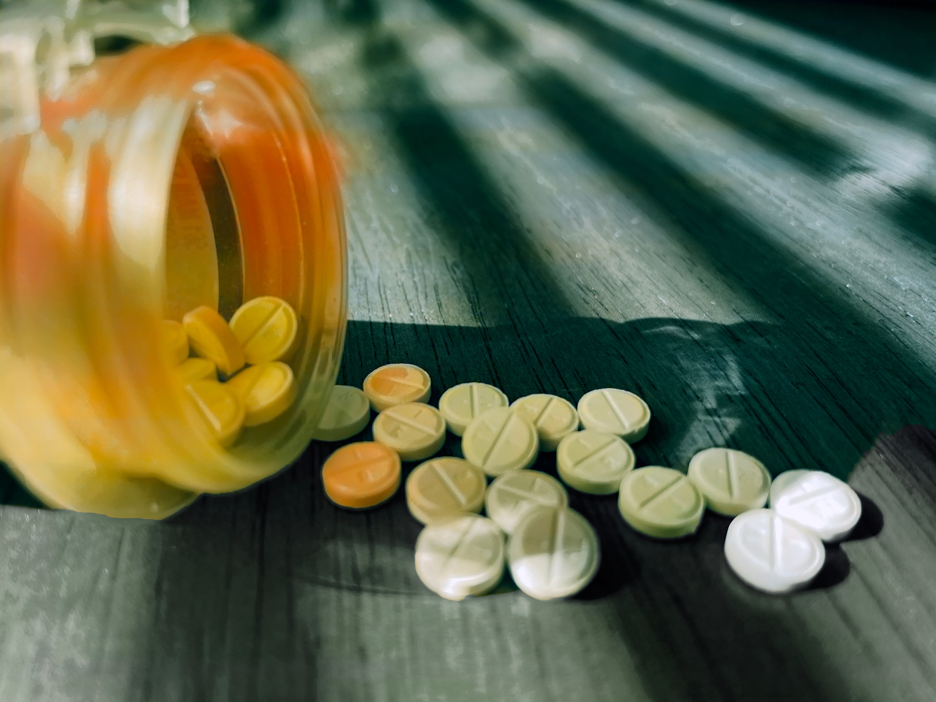 Pills scattered from a tipped-over bottle on a wooden surface, casting shadows from blinds