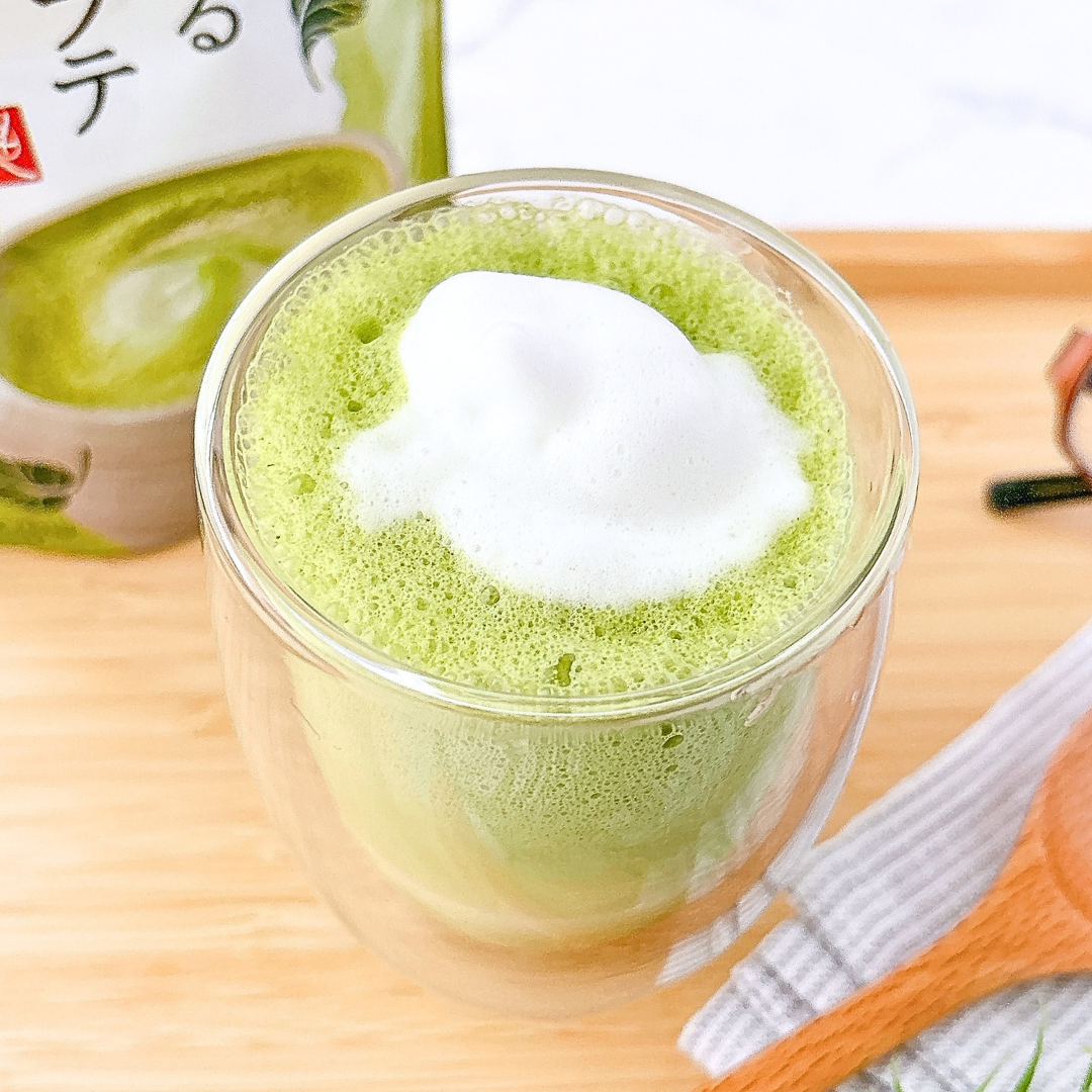 泡がのった抹茶ラテの入った透明なコップと木のスプーン。背景に抹茶のパッケージが見える。