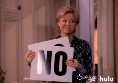 Person holding a sign with "NO" printed on it, giving an amused expression in a domestic setting Scene from "Sabrina" series