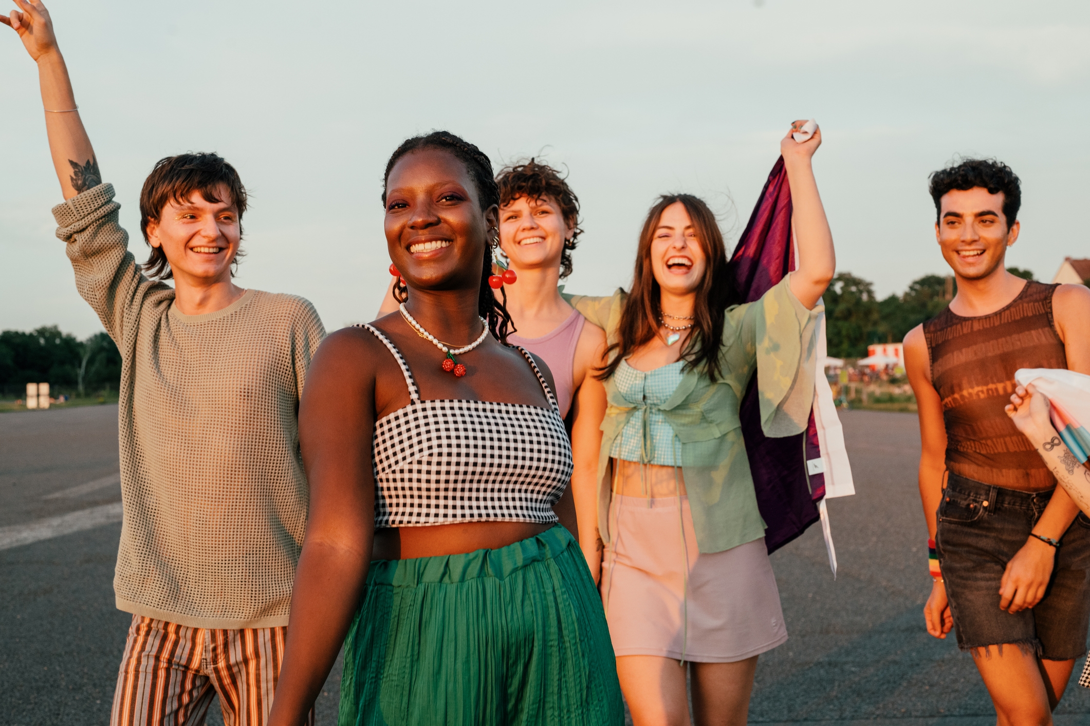 Group of five people outdoors, smiling and walking. One person raises an arm triumphantly, while another holds a flag. Casual, summery attire