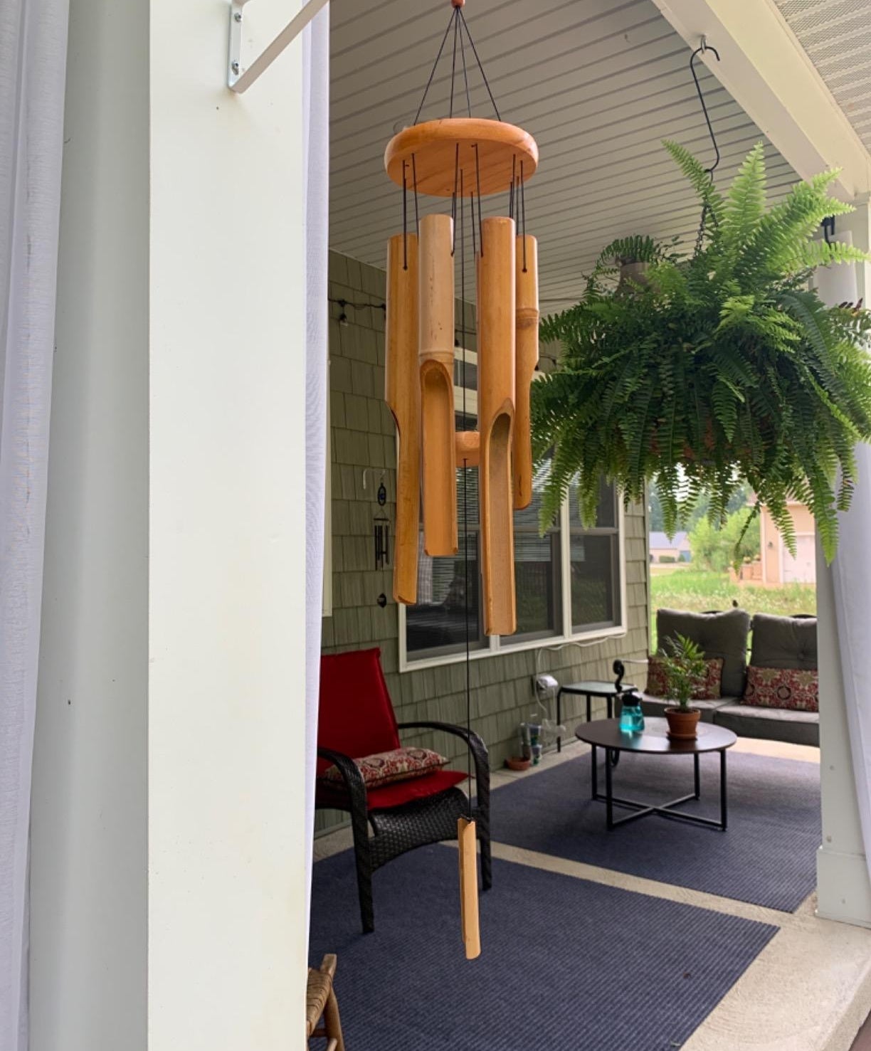 A porch with a hanging wooden wind chime and a large fern. There is outdoor seating with cushions and a small table with a bottle on it