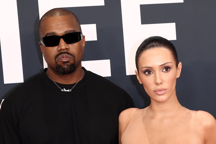 Two individuals posing on a red carpet; person on the left wears sunglasses and a dark top, person on the right wears a strapless outfit