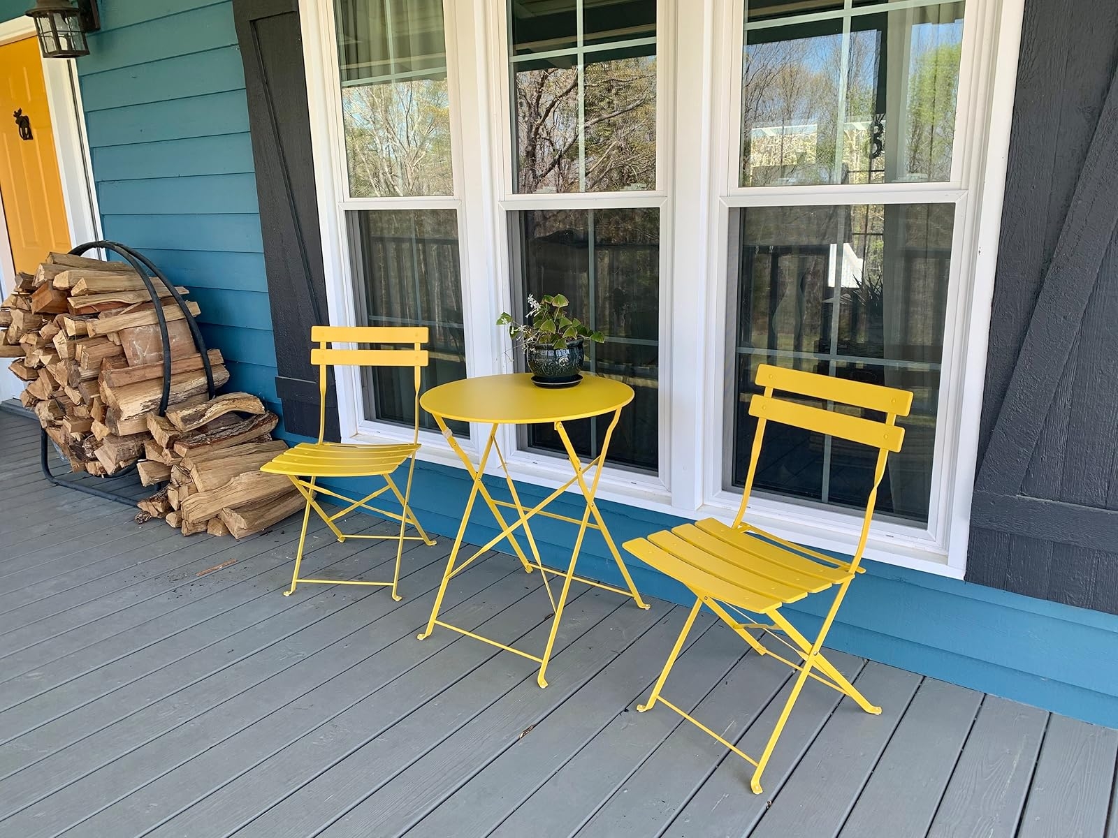 Two yellow chairs and a round table with a plant sit on a porch with stacked firewood beside a window