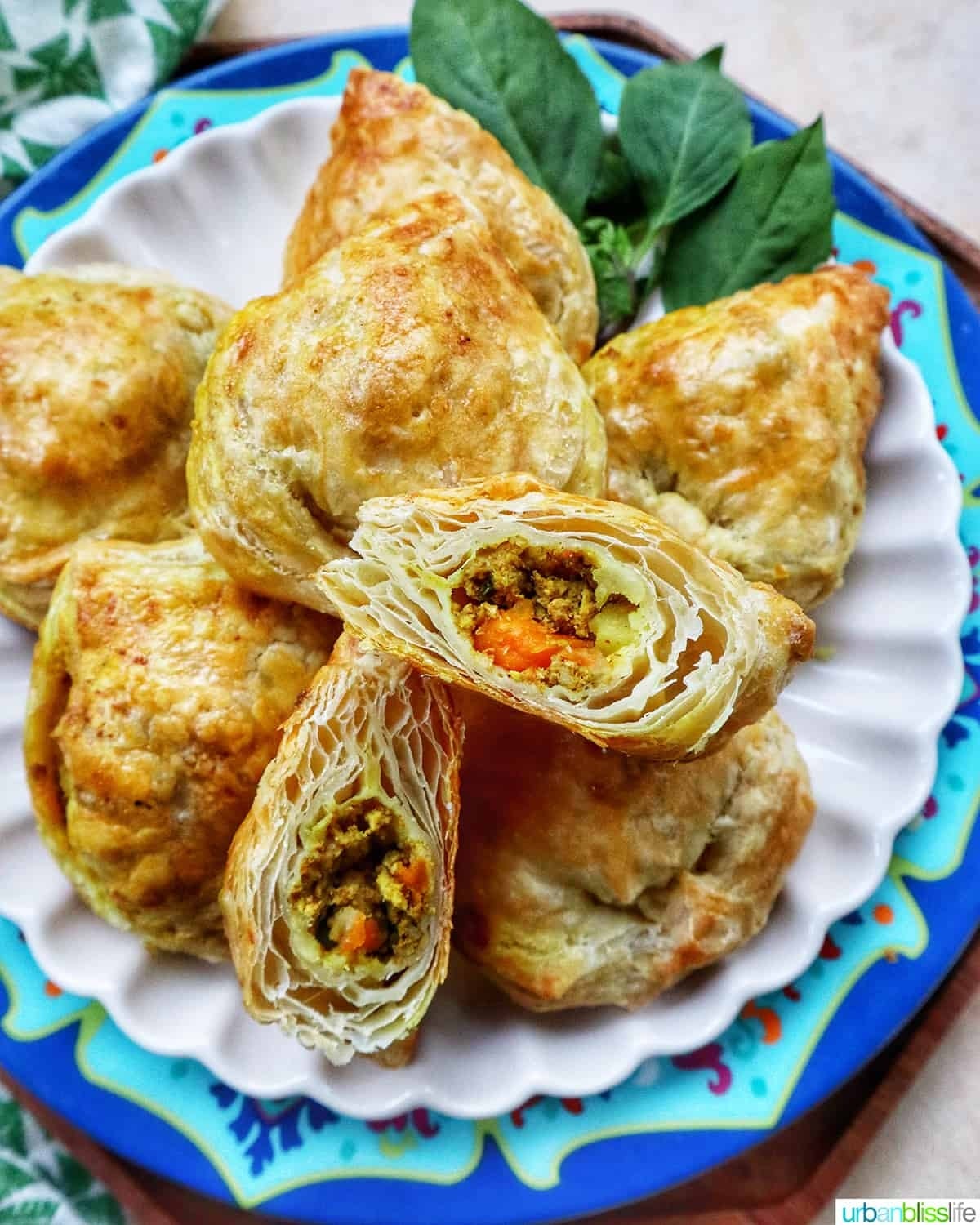A plate of flaky, golden-brown curry puffs filled with a savory mixture of vegetables and spices, arranged decoratively with fresh basil leaves