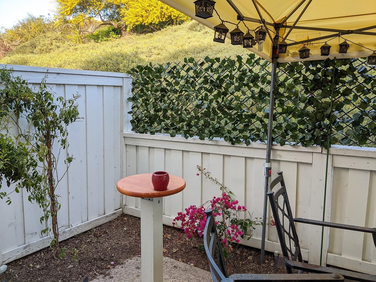 A cozy backyard corner with a small round table and a decorative chair, surrounded by a white fence and greenery, under a canopy