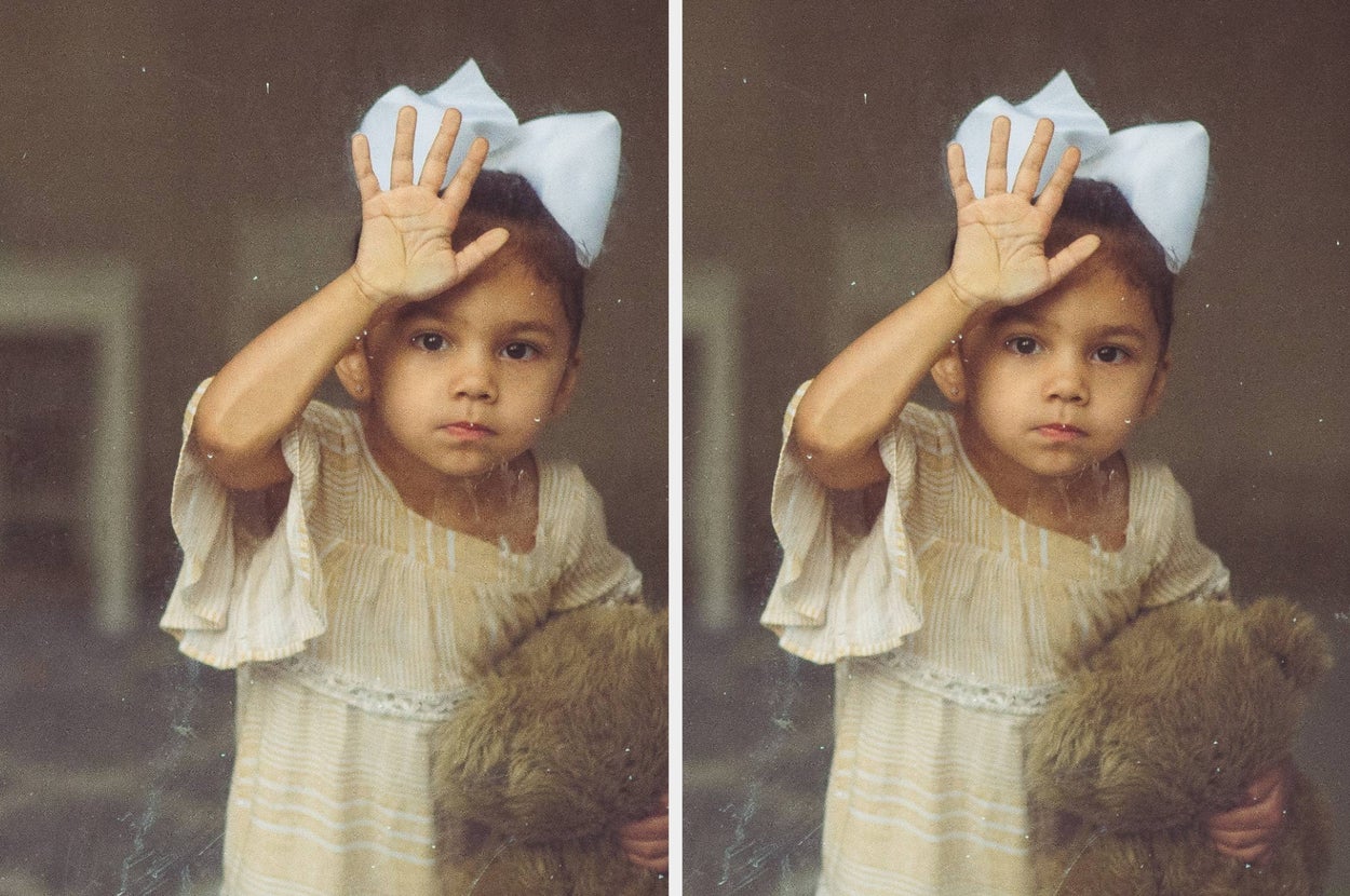 Child with a bow in hair touches window, holding teddy bear, gaze serious