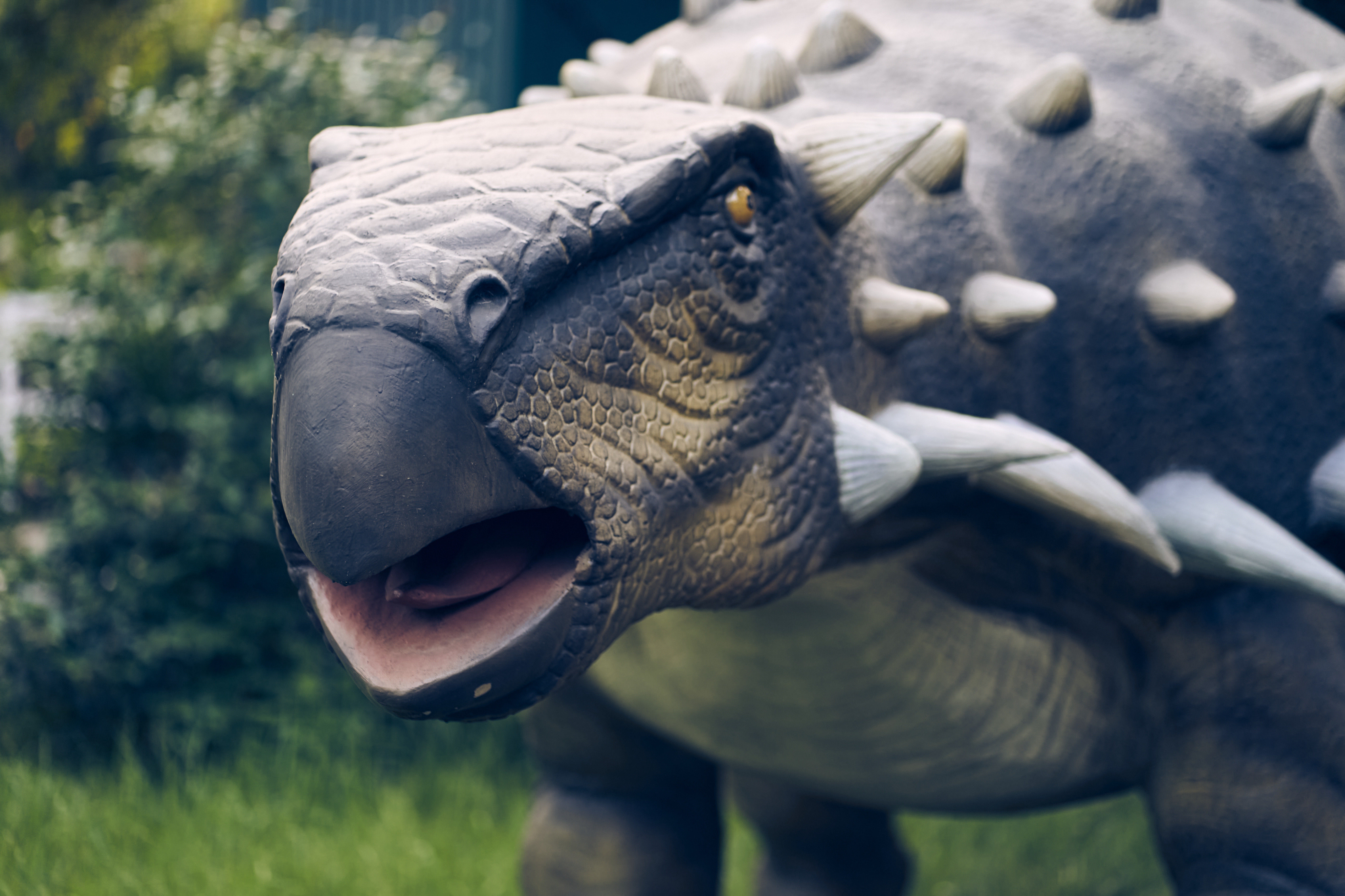 A realistic dinosaur model with textured skin and spiked armor stands outdoors, set against a blurred park background