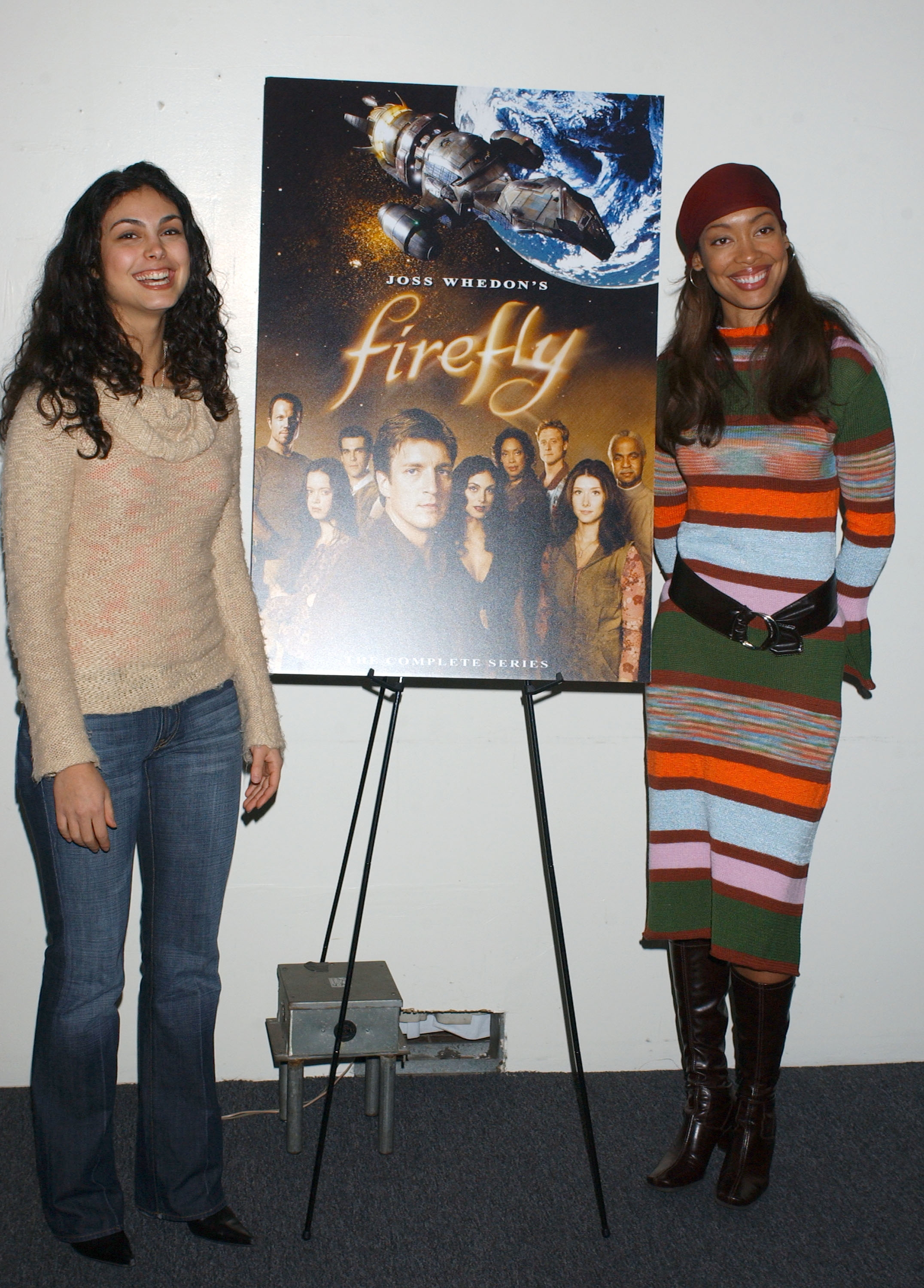 Morena Baccarin and Gina Torres stand beside a "Firefly" poster. The woman on the left wears a sweater and jeans, the woman on the right is in a striped dress and boots
