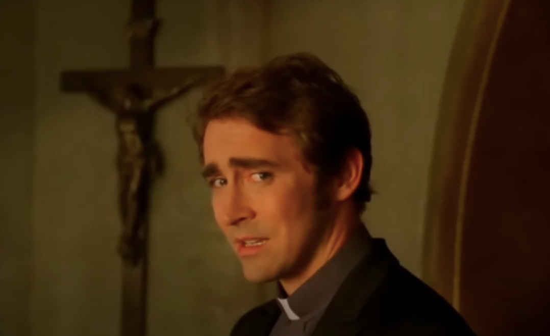 Lee Pace in a clerical collar looks over his shoulder with a concerned expression, standing in front of a large crucifix