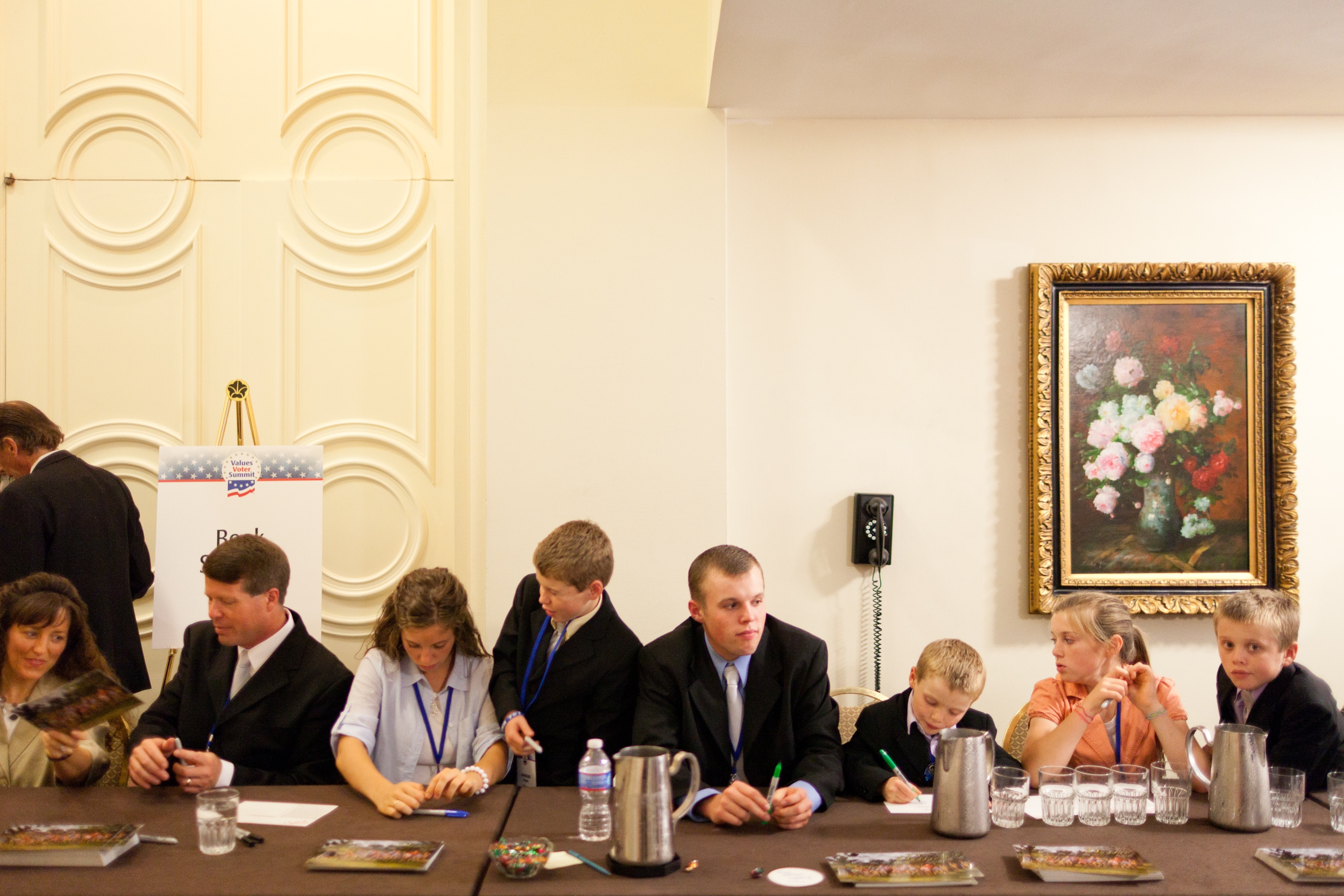 The Duggar family seated at a long table reviewing documents. Walls have classic decor and a floral painting