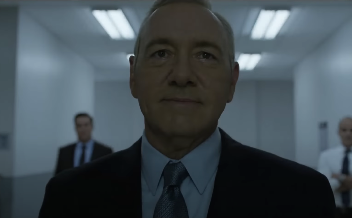 Kevin Spacey in a suit walks confidently down a brightly lit hallway, with two blurred figures in the background