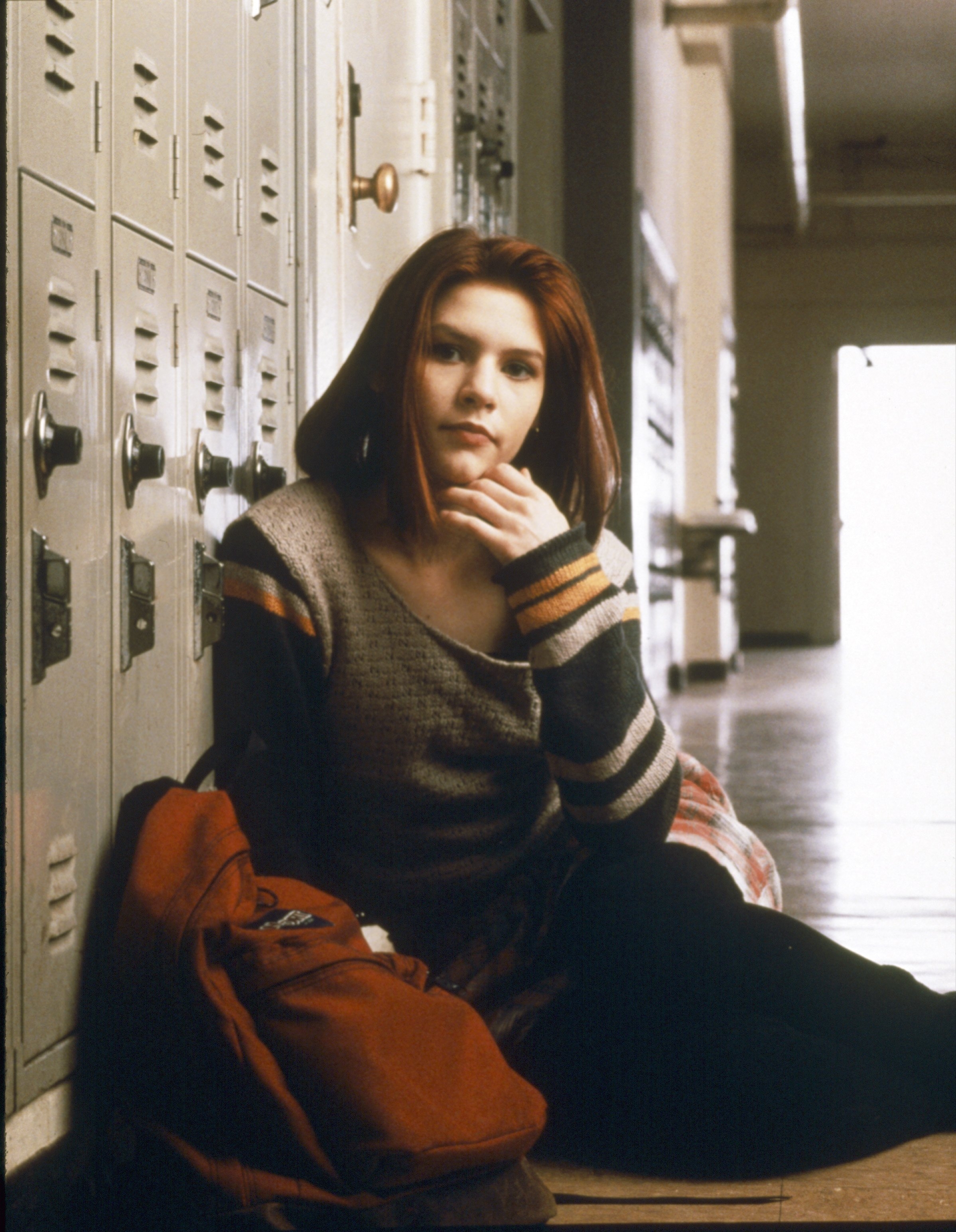 Clare Danes on the floor by lockers, wearing a casual sweater. They have an orange backpack beside them and appear contemplative