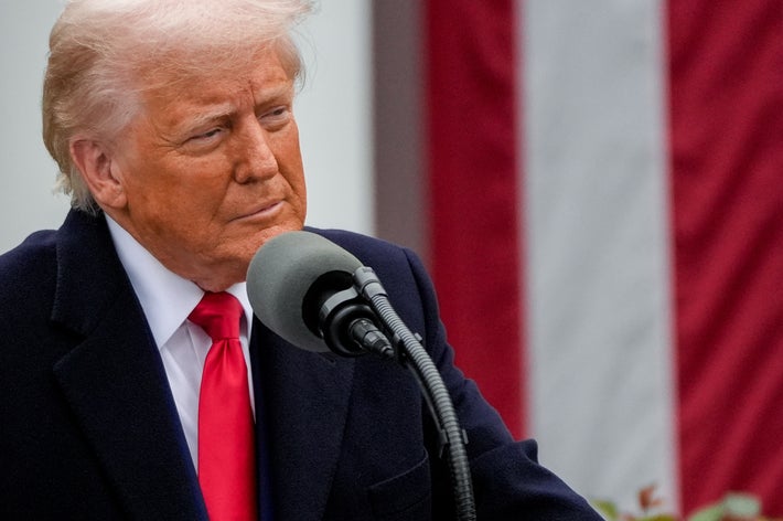 男性が屋外でマイクに向かってスピーチをしている。背景に縞模様の旗が見える。