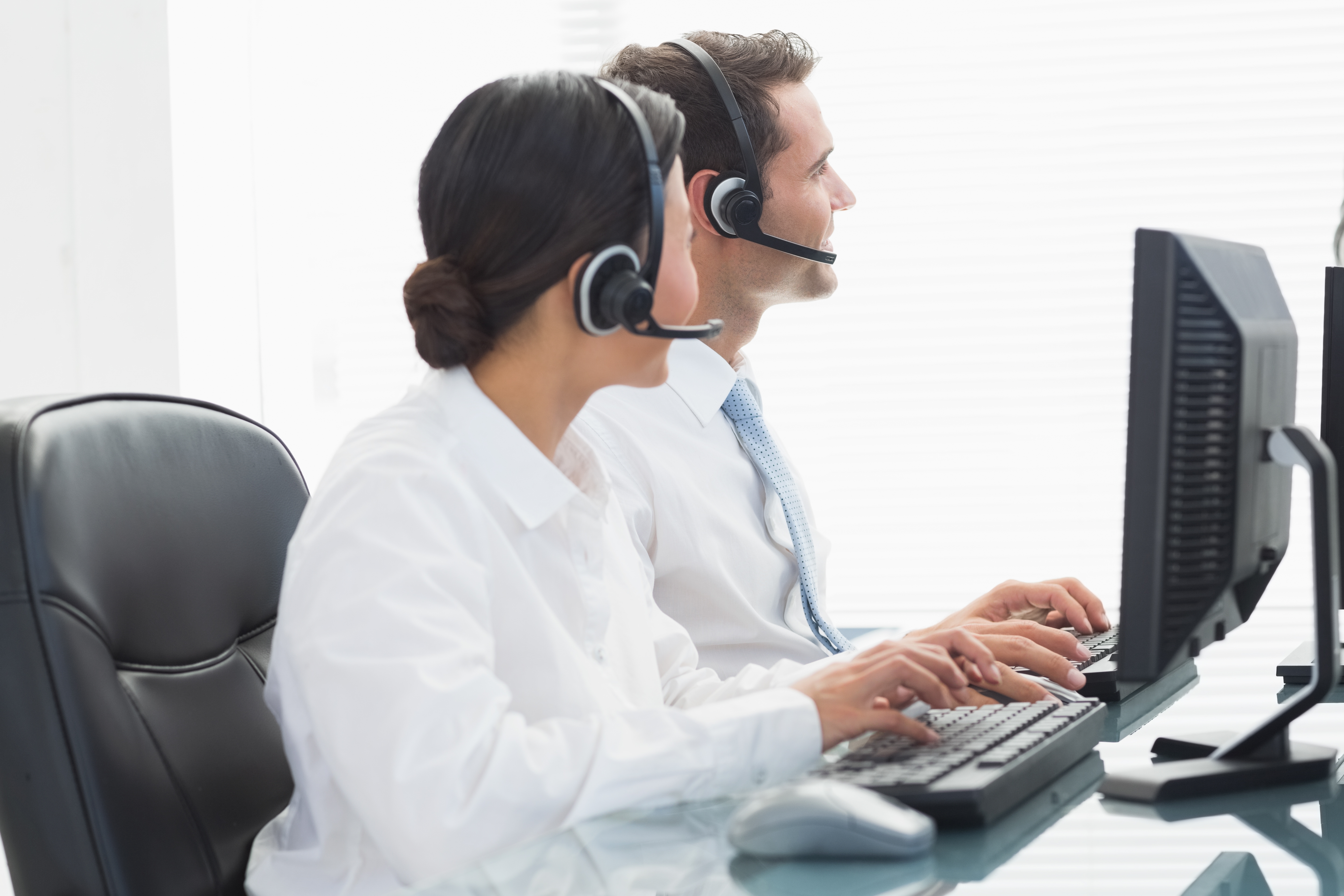 Two people in headsets, sitting at desks with computers, focus on their screens. Scene depicts a busy office work environment