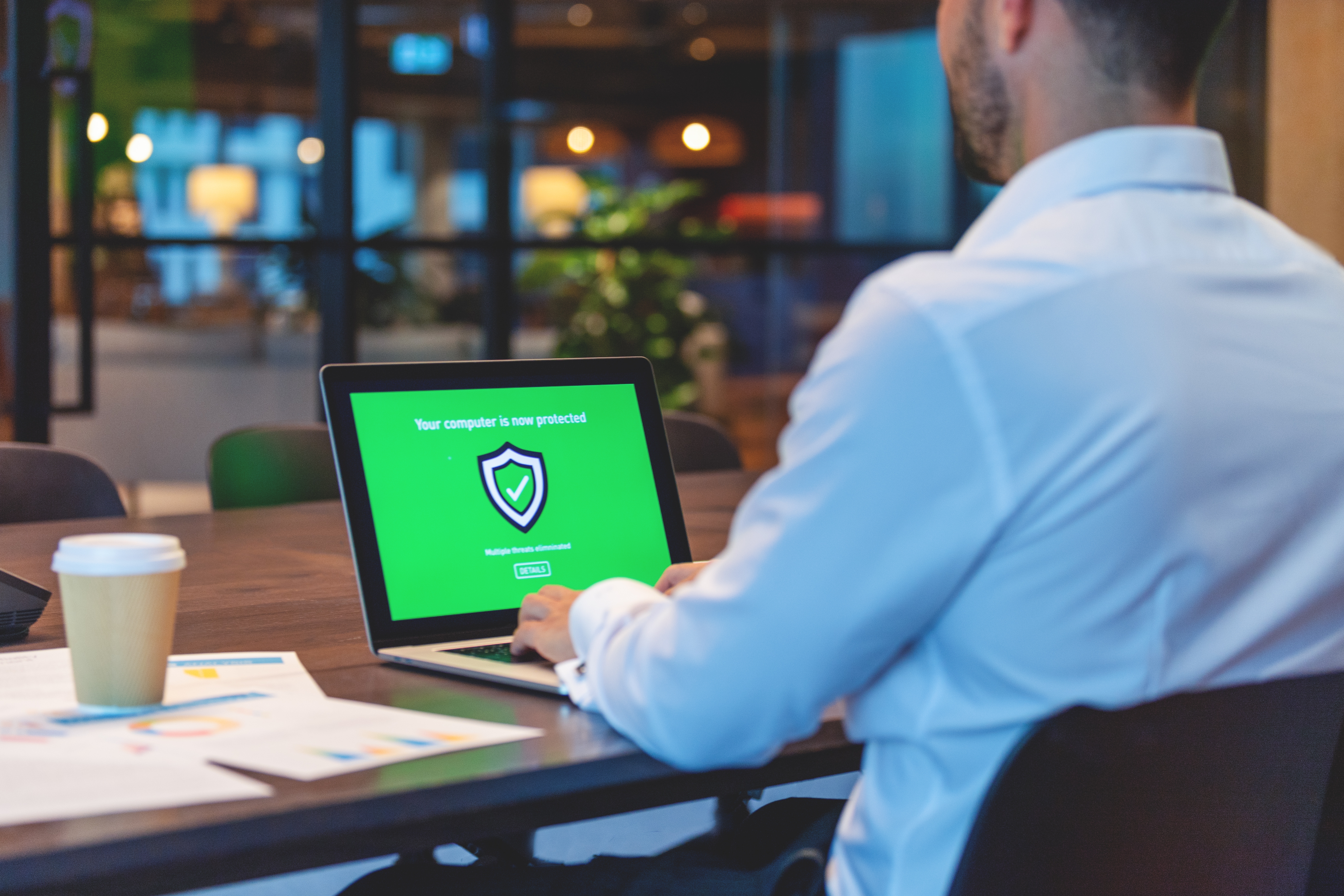 Man at a desk using a laptop with a green antivirus protection screen. A coffee cup and documents are nearby, suggesting a work setting