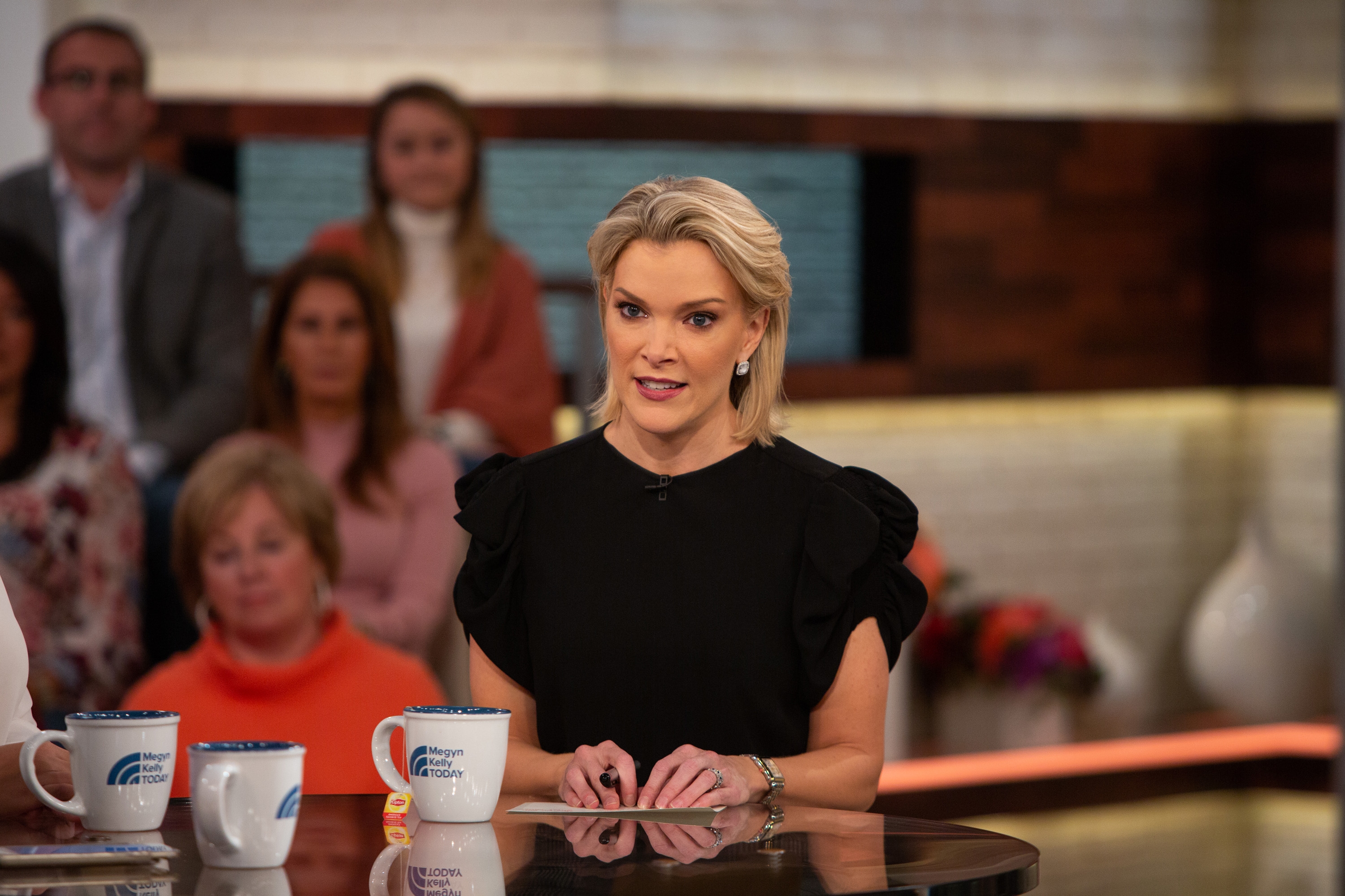 Megyn Kelly in a TV studio setting, wearing a ruffled black blouse, appears to be hosting a discussion panel with an audience in the background