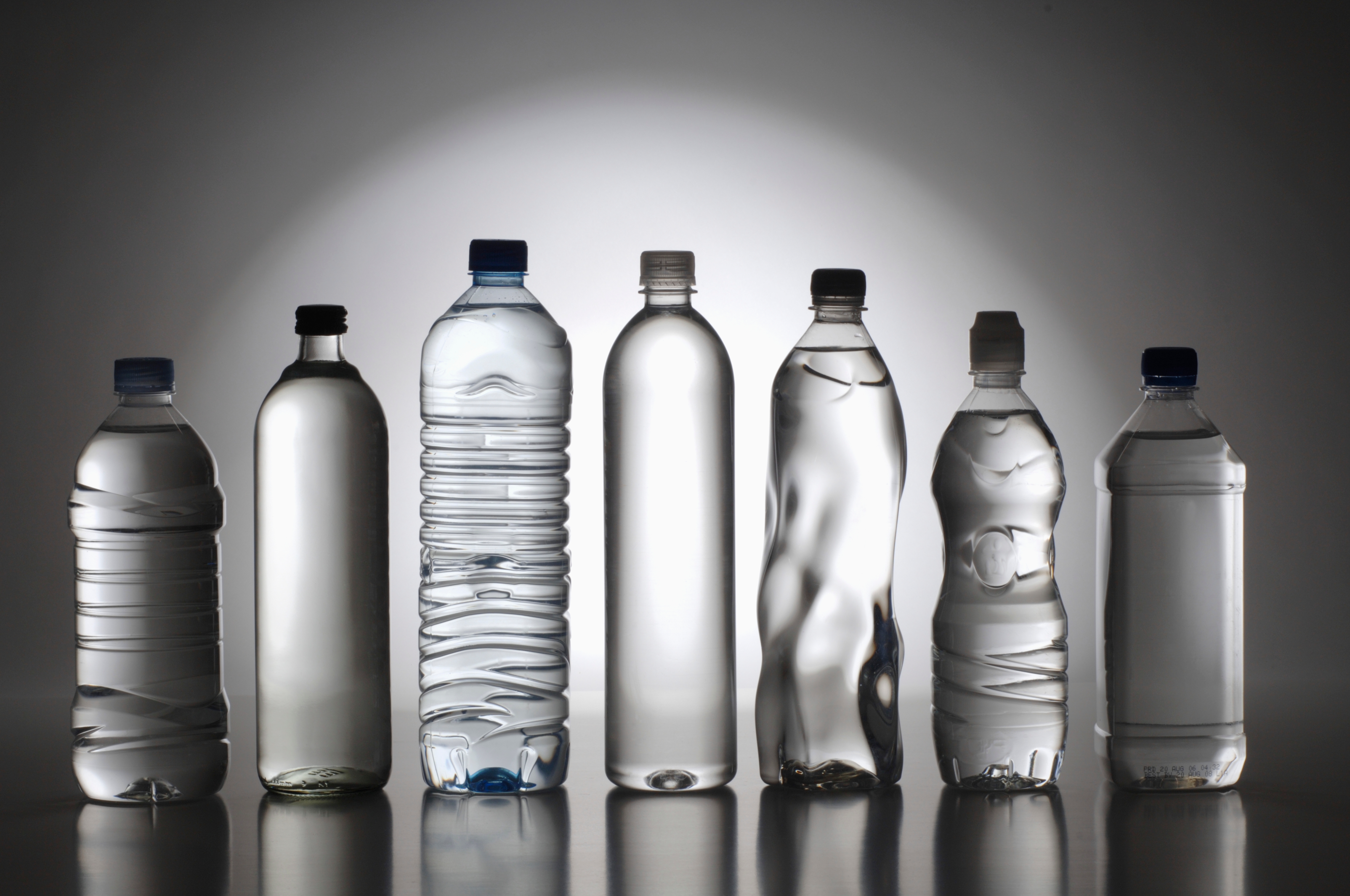 Seven different types of plastic water bottles are lined up in a row, showcasing variety in design and shape, relevant to a discussion on consumer goods