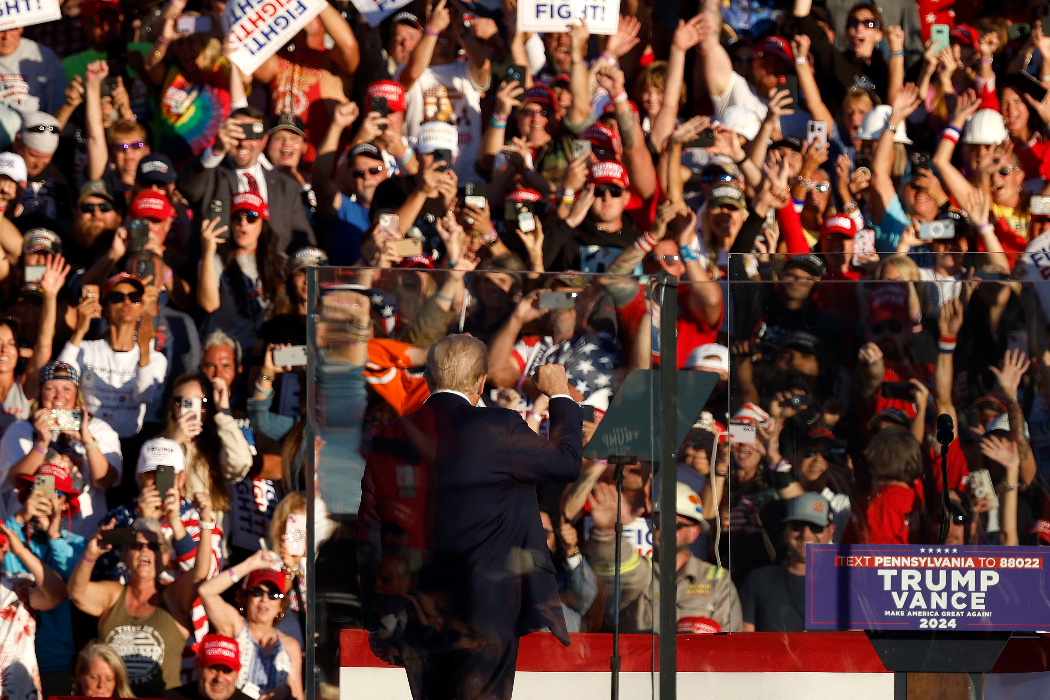 A political figure gestures to a large cheering crowd at a rally. A "Text Pennsylvania to 88022 Trump Vance 2024" sign is visible