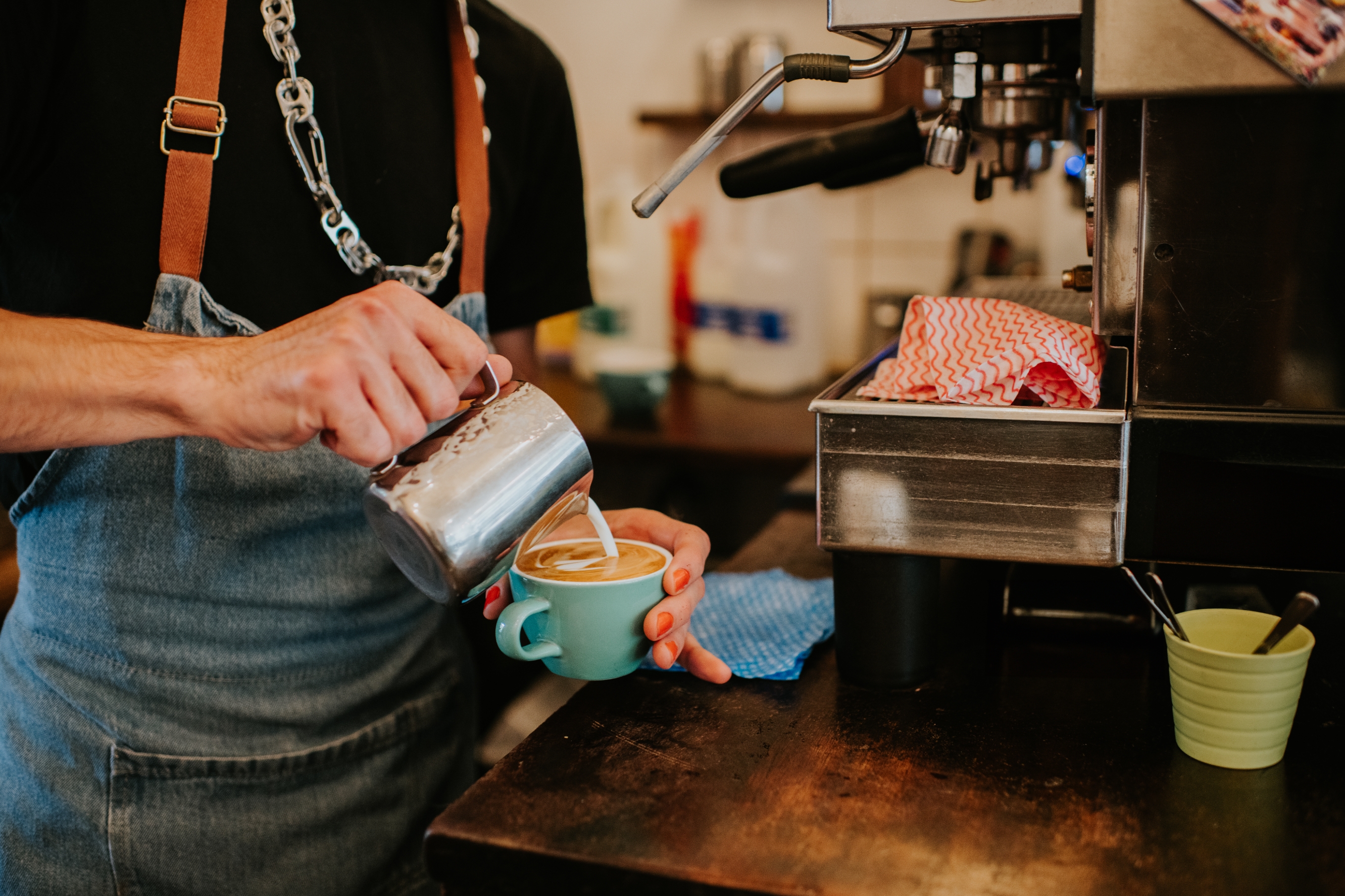 Barista in apron and chain necklace pouring latte art in a café, highlighting artistic coffee-making skills