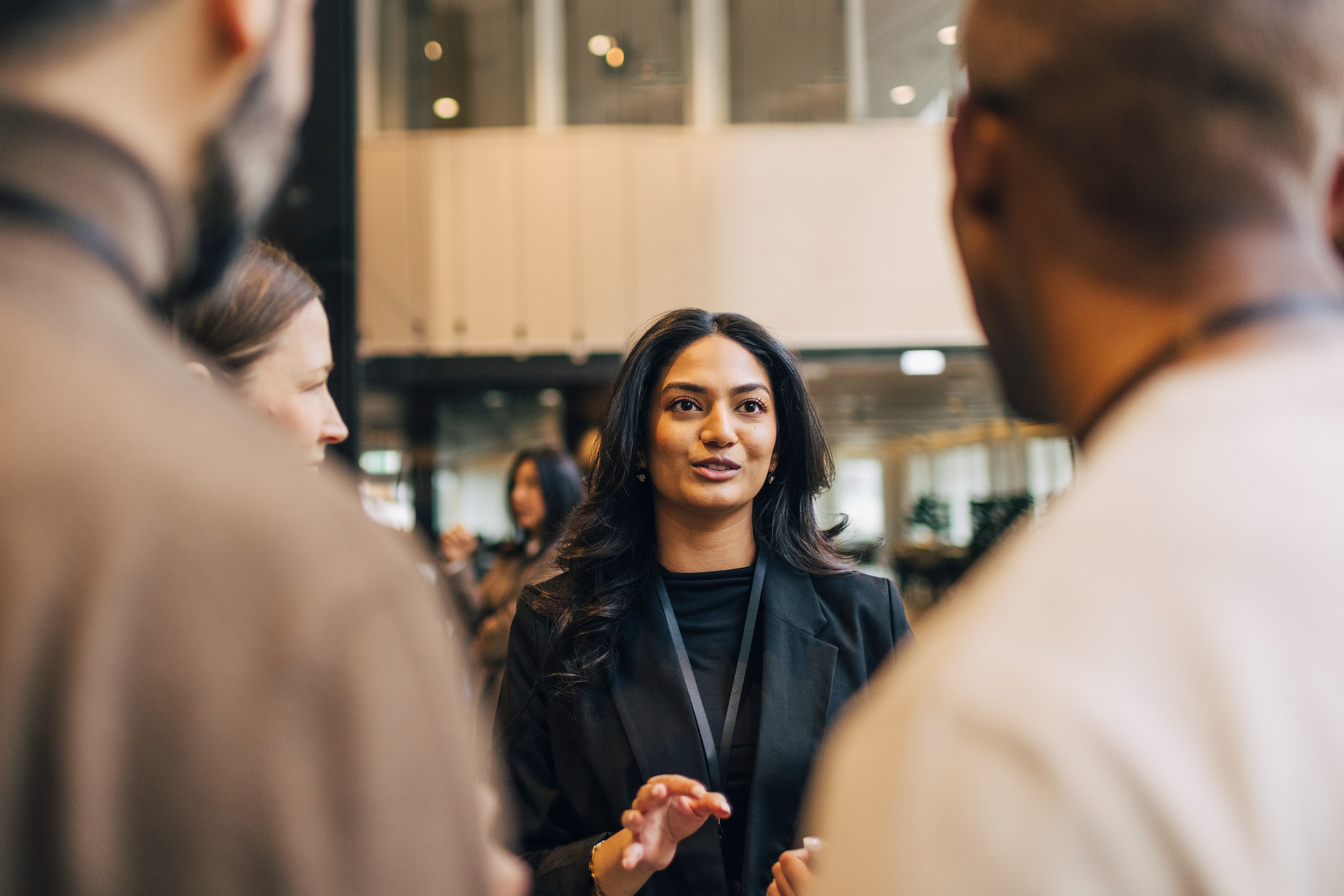 Person in a business setting speaking to colleagues, wearing a professional blazer. The focus is on work collaboration and communication
