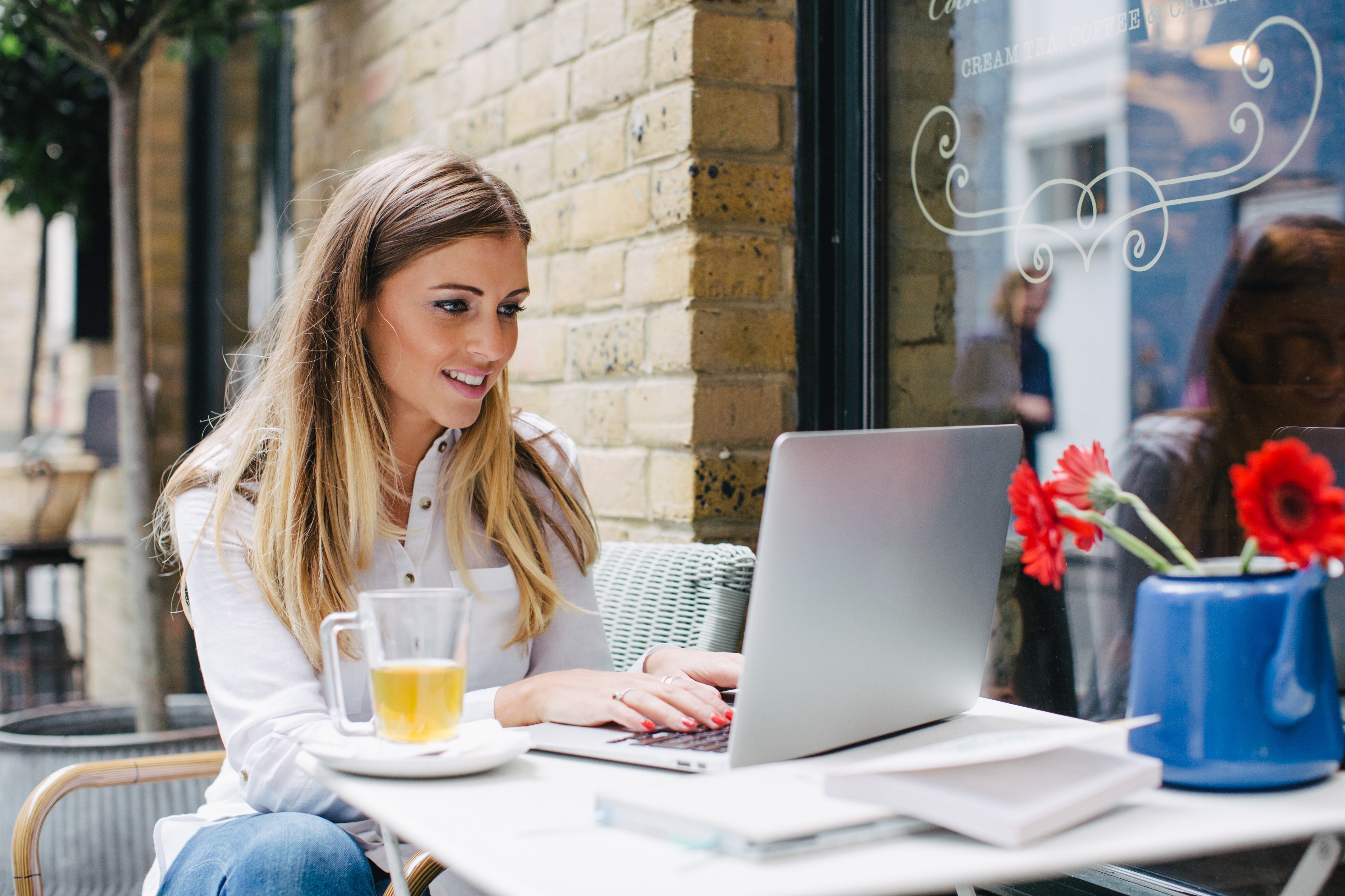 Woman in a shirt works on a laptop at a cafe table, with a glass mug of tea and a vase with red flowers beside her