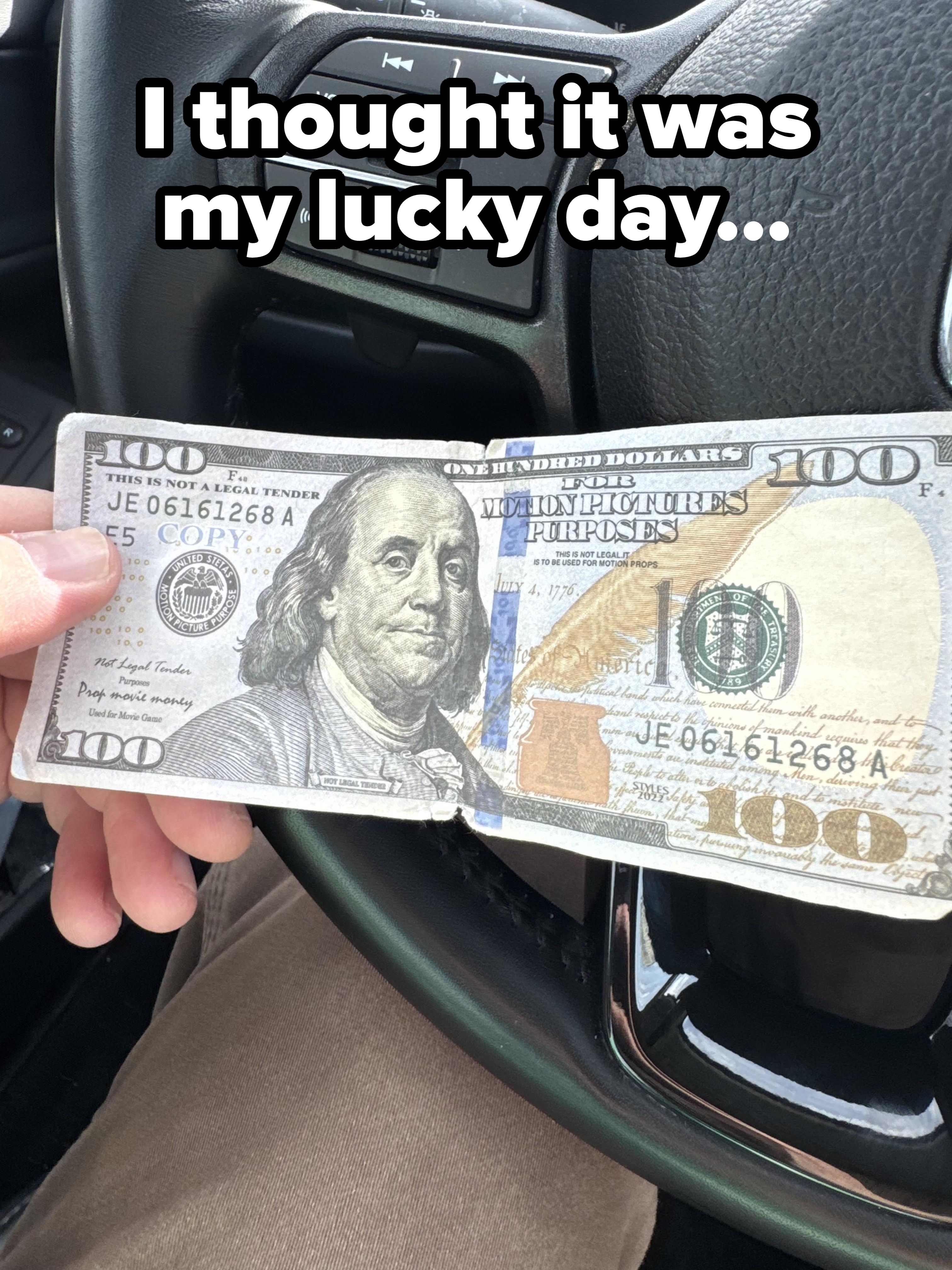 Person holding a folded $100 bill inside a car, with a steering wheel and controls visible