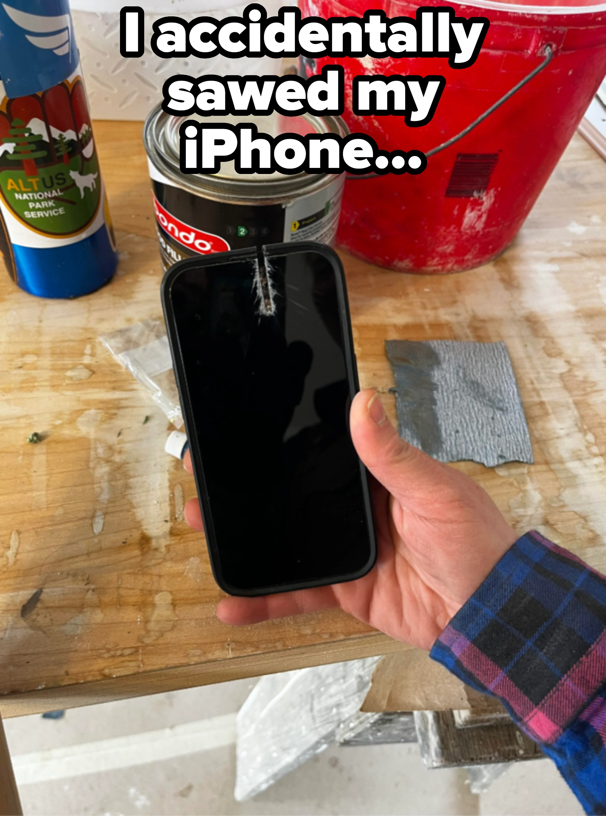 Person holding a smartphone with a cracked screen on a workbench, surrounded by tools and paint cans