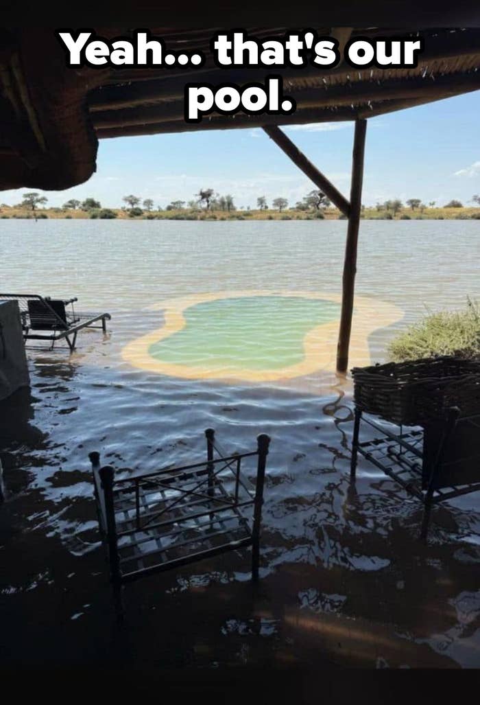 Chairs and a small table partially submerged in water by a riverside, under a canopy, with a view of water and distant trees
