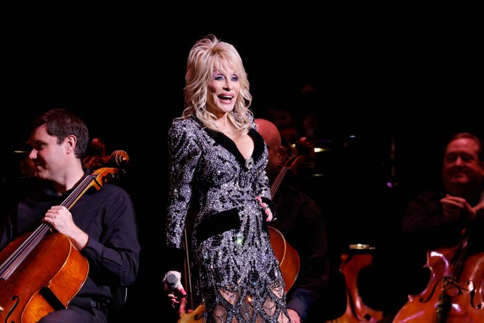 A singer in a sparkling, form-fitting dress performs on stage with an orchestra, holding a microphone and smiling