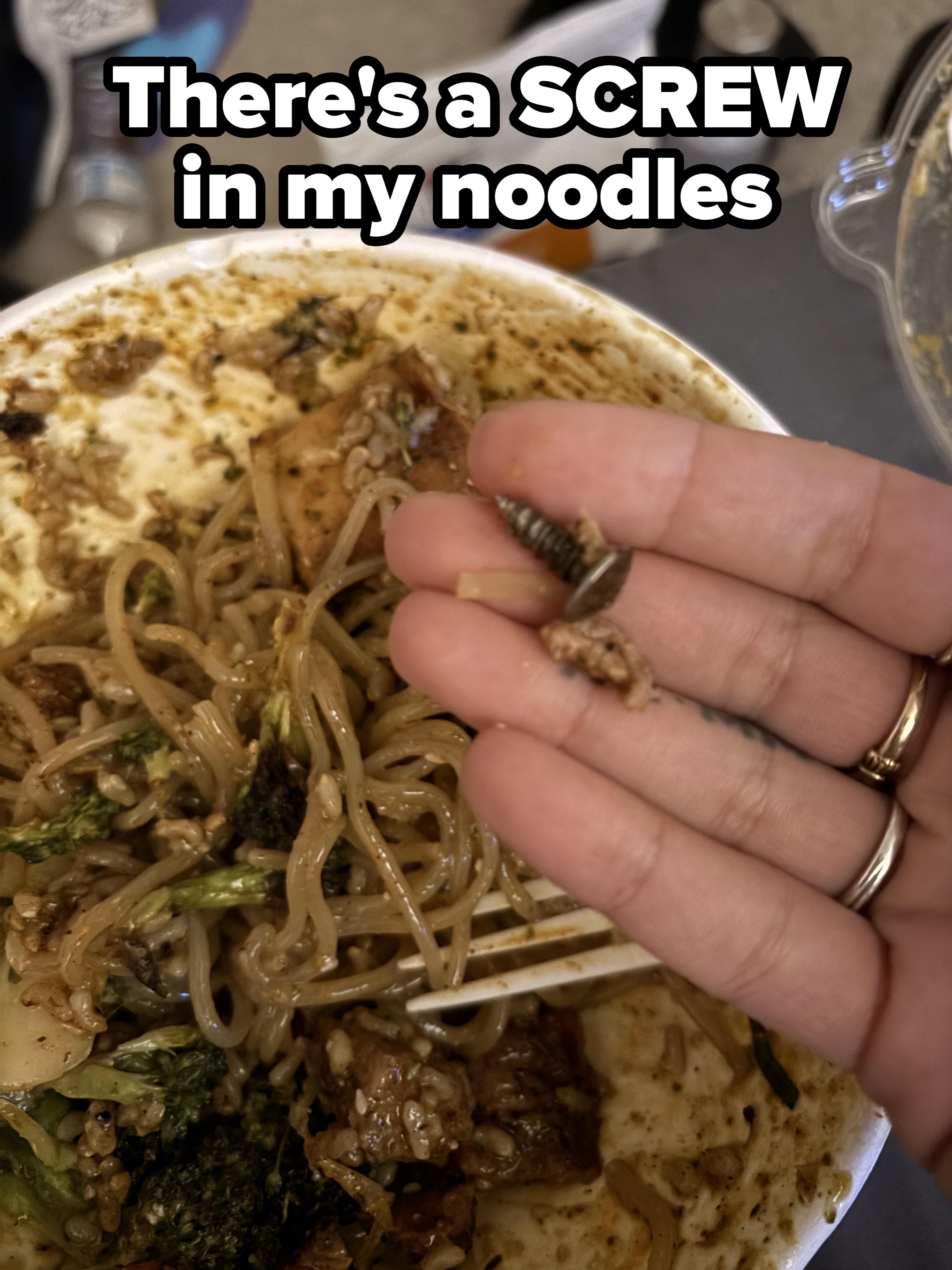 Person holding a screw found in a bowl of noodles with vegetables, capturing a surprising dining mishap