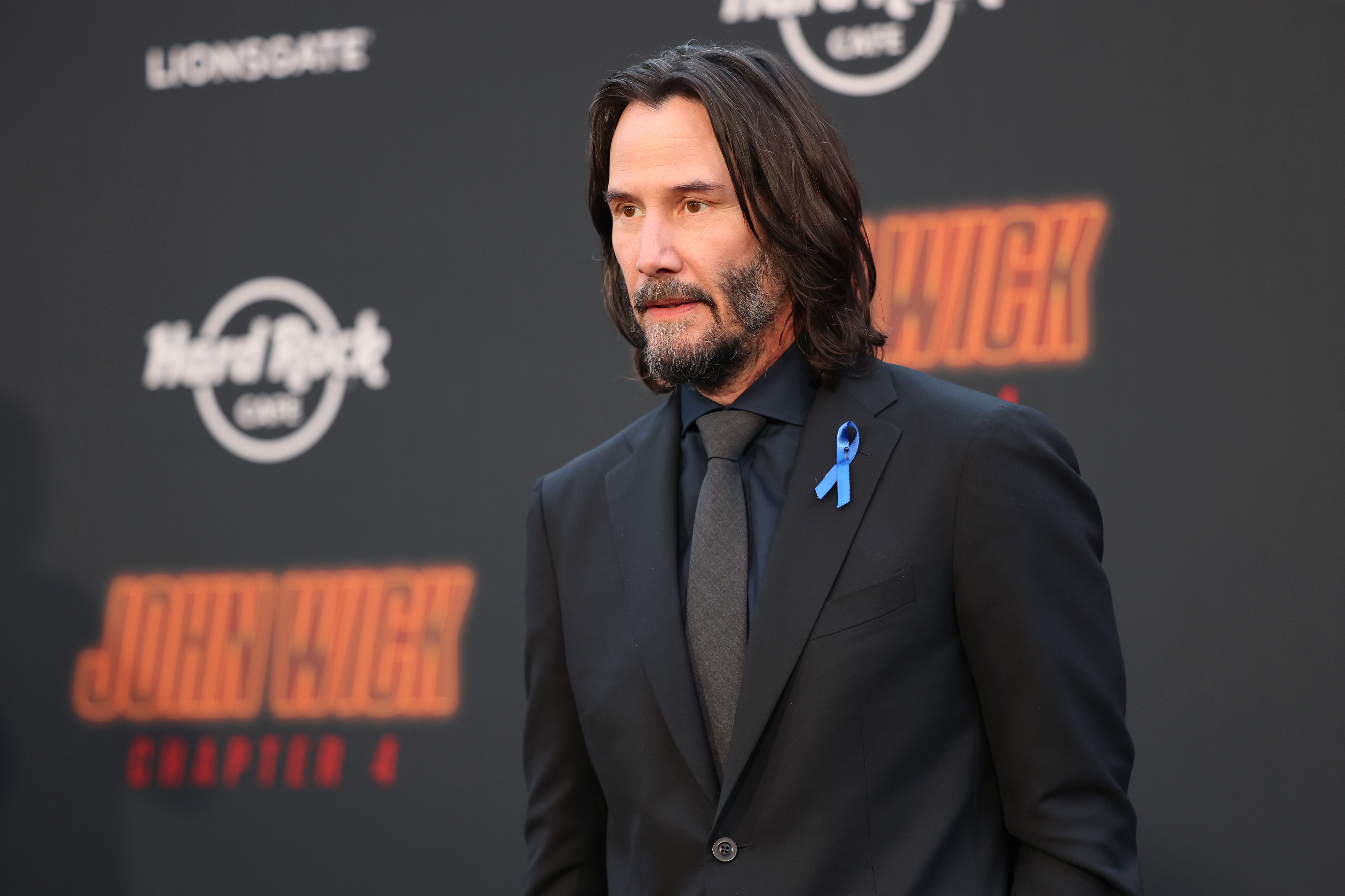 A person in a black suit with a blue awareness ribbon is on a red carpet at a &quot;John Wick: Chapter 4&quot; event