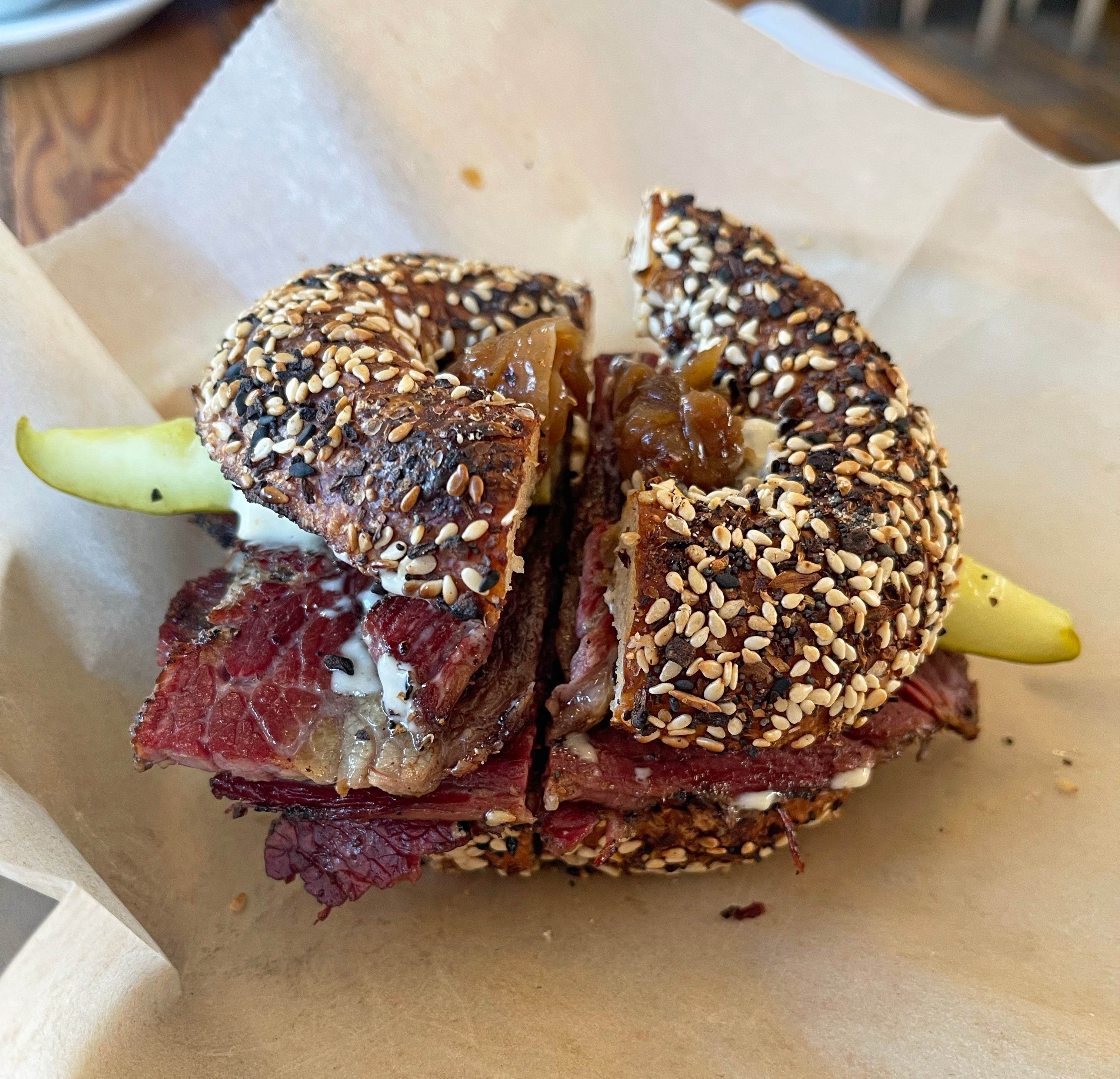 Everything bagel sandwich with pastrami, cream cheese, caramelized onions, and a pickle slice on parchment paper