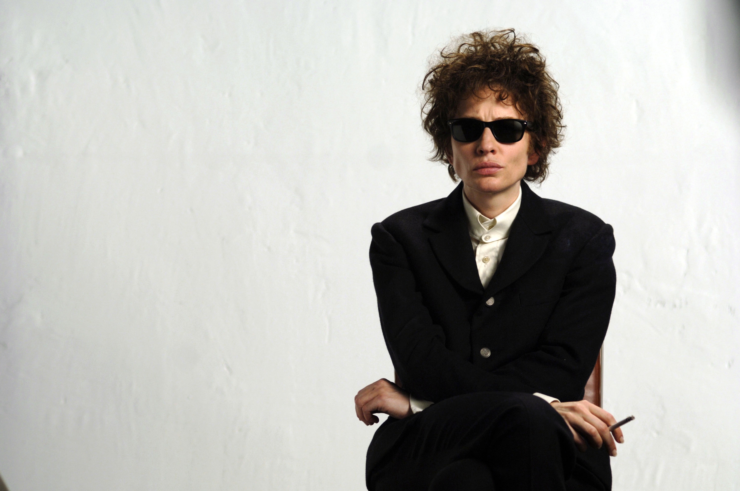 Cate Blanchett as Bob Dylan in "I'm Not There" sat on a chair, arms crossed over lap, holding a cigarette and wearing sunglasses