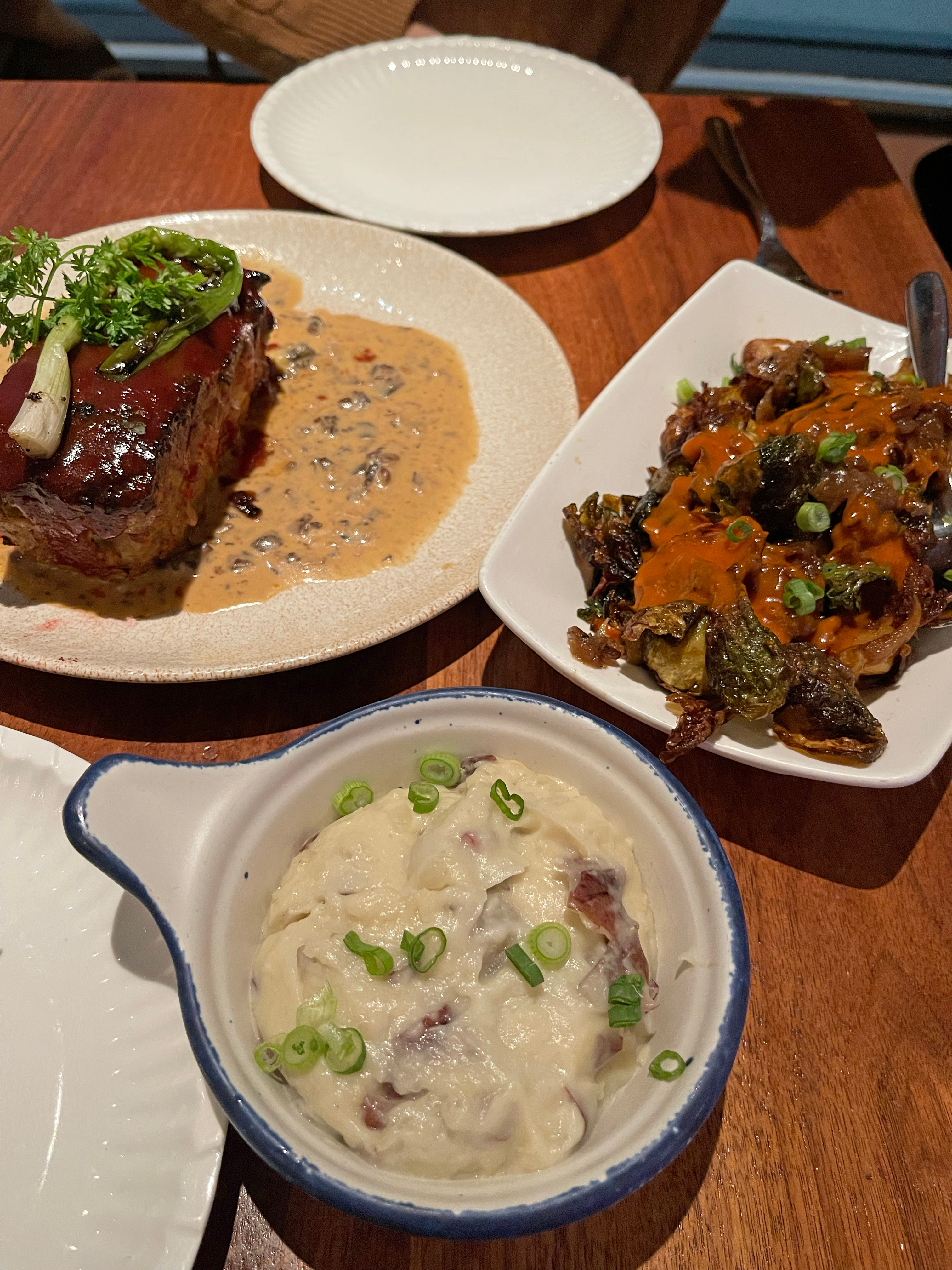 A plate with steak and sauce, a dish of roasted Brussels sprouts with orange sauce, and a bowl of mashed potatoes with green onions on a wooden table