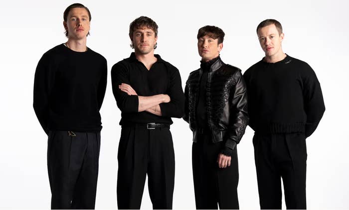 Four individuals stand posed, wearing stylish black outfits; one wears a leather jacket. They all face the camera with neutral expressions