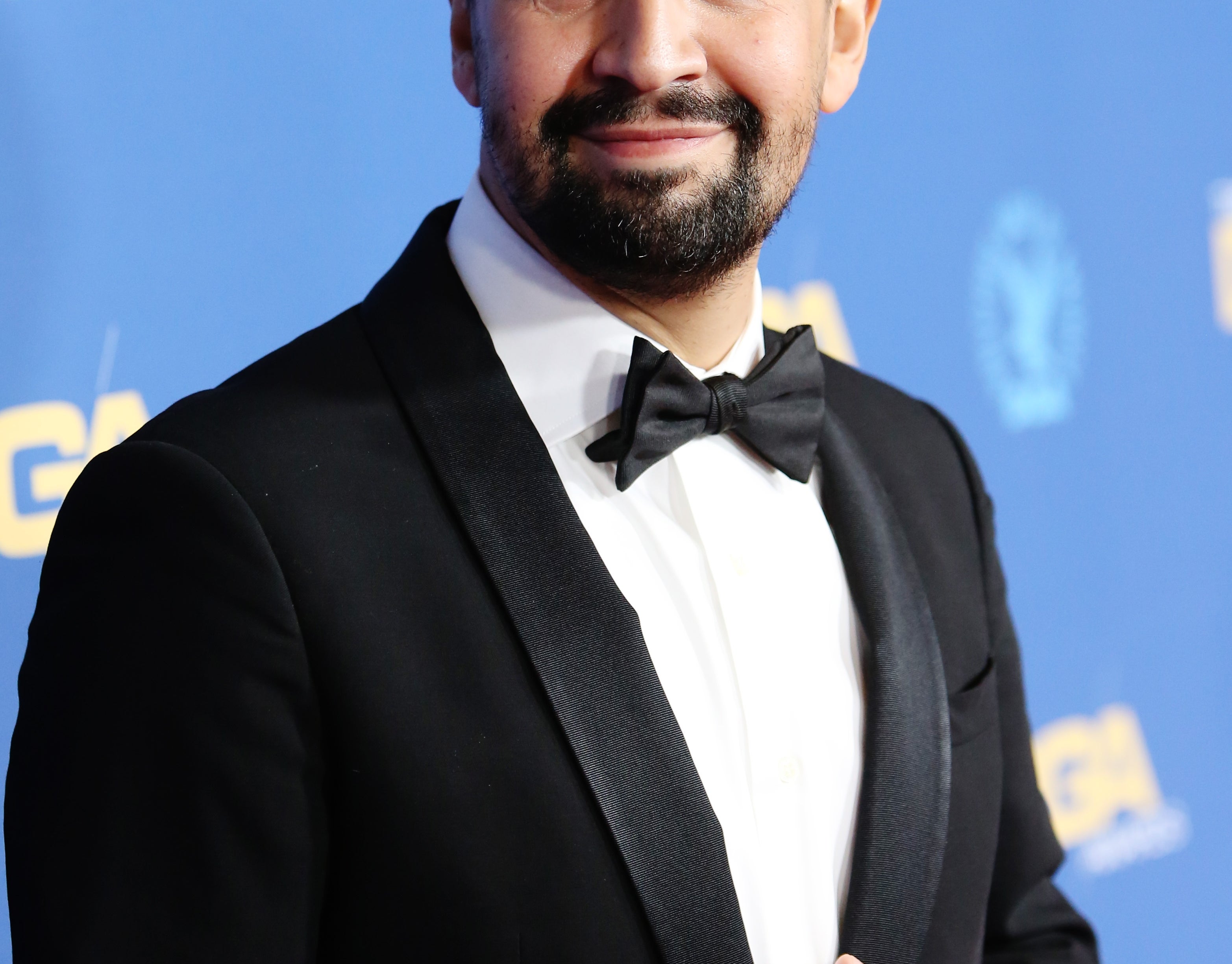 Person in a tuxedo with a bow tie on a blue backdrop at a formal event, smiling and looking to the side