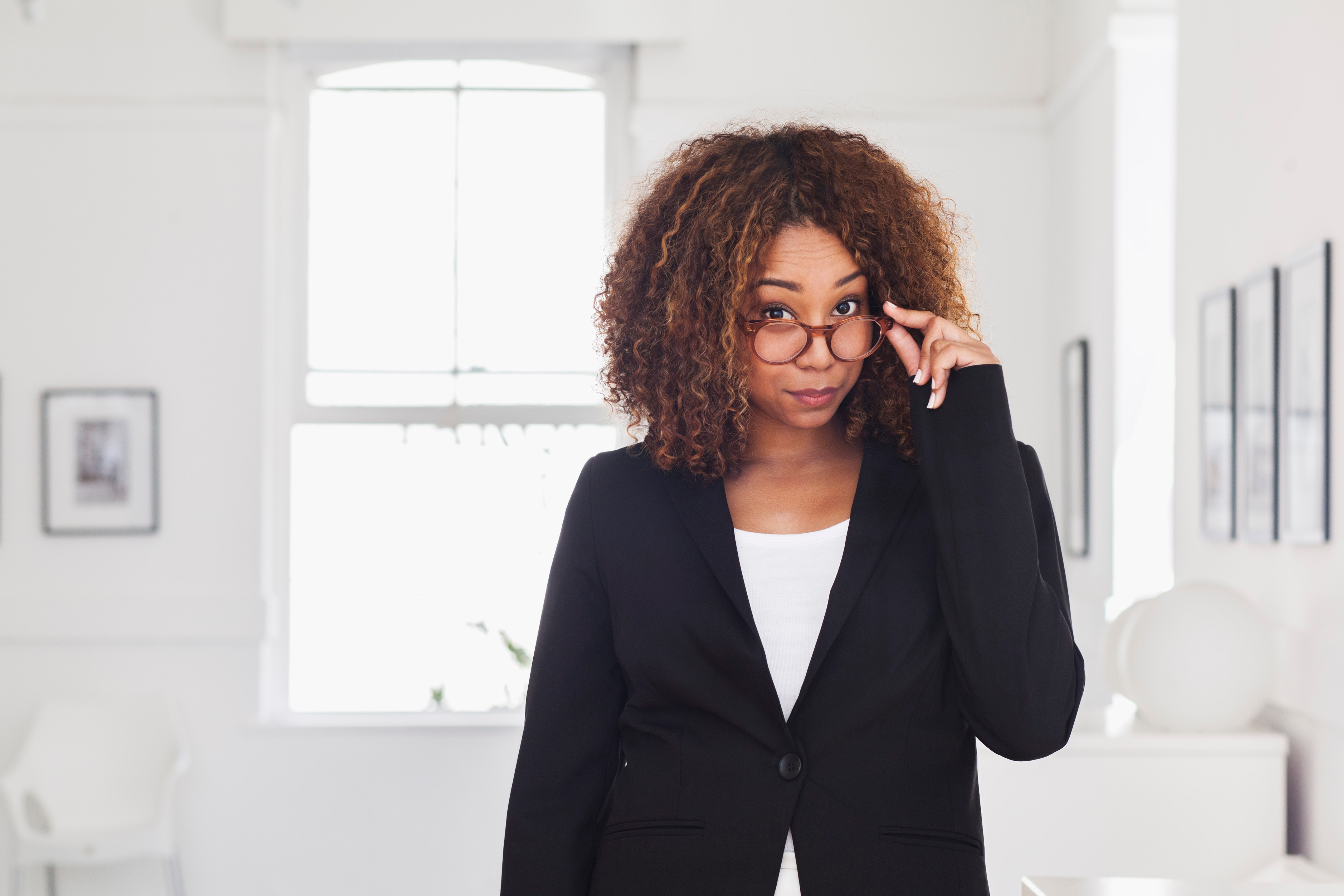 Person with curly hair wearing glasses and a blazer, standing indoors, looking over the glasses with a confident expression. Indoors setting visible