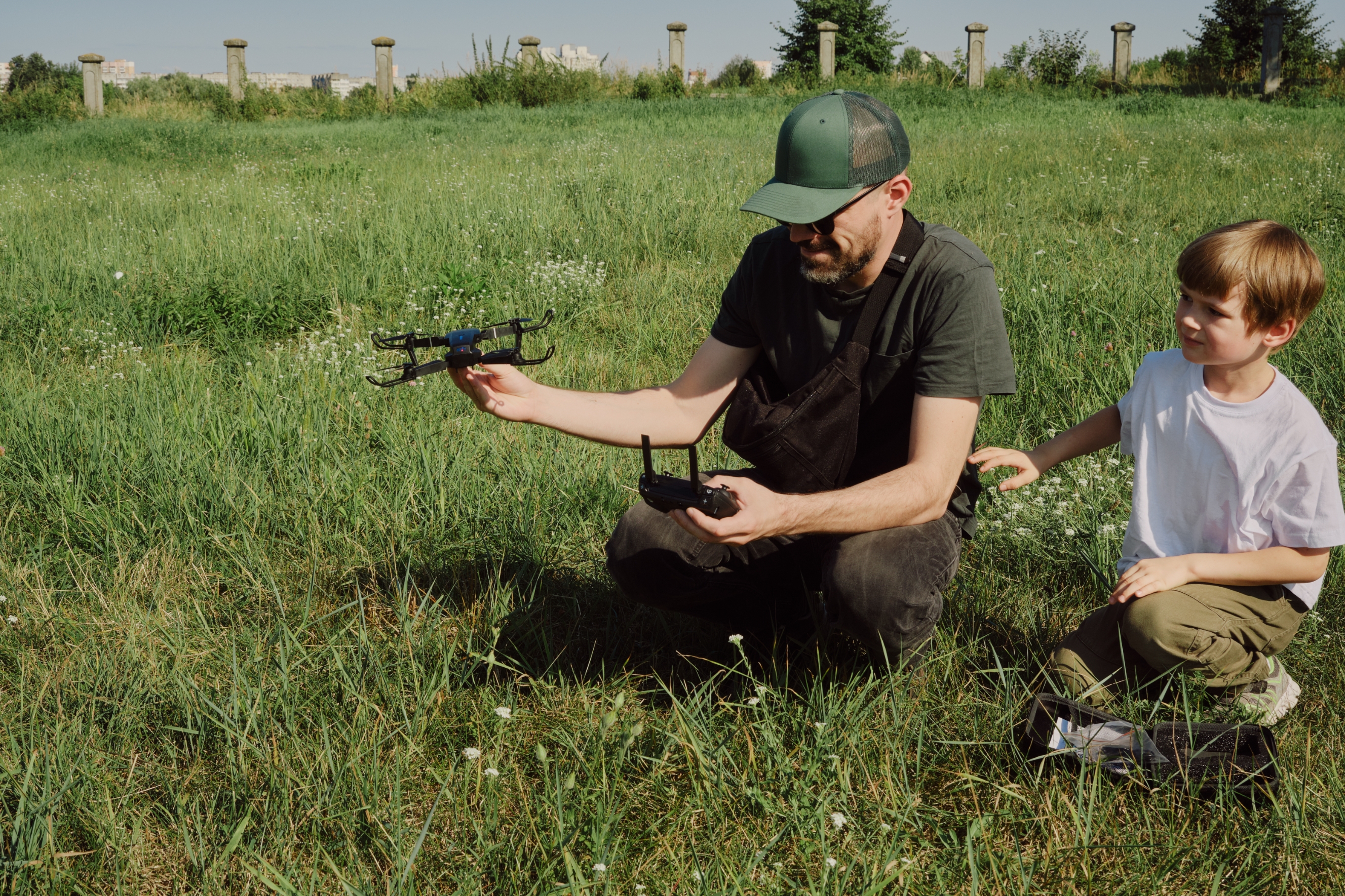 A man kneeling in a grassy field demonstrates a drone to a young boy watching attentively