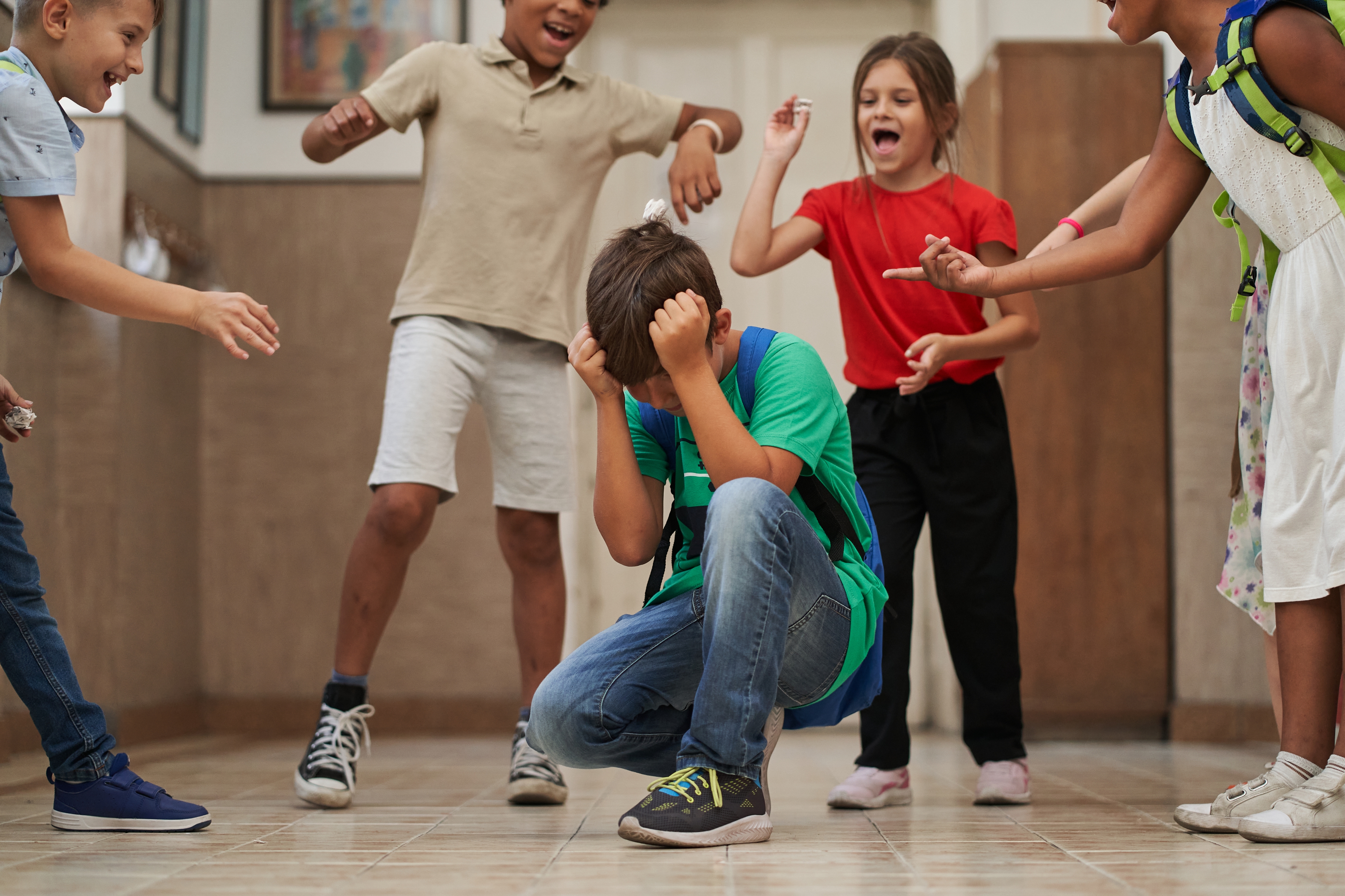 Children surround and tease a boy crouched on the floor in a hallway, highlighting a scene of bullying and distress
