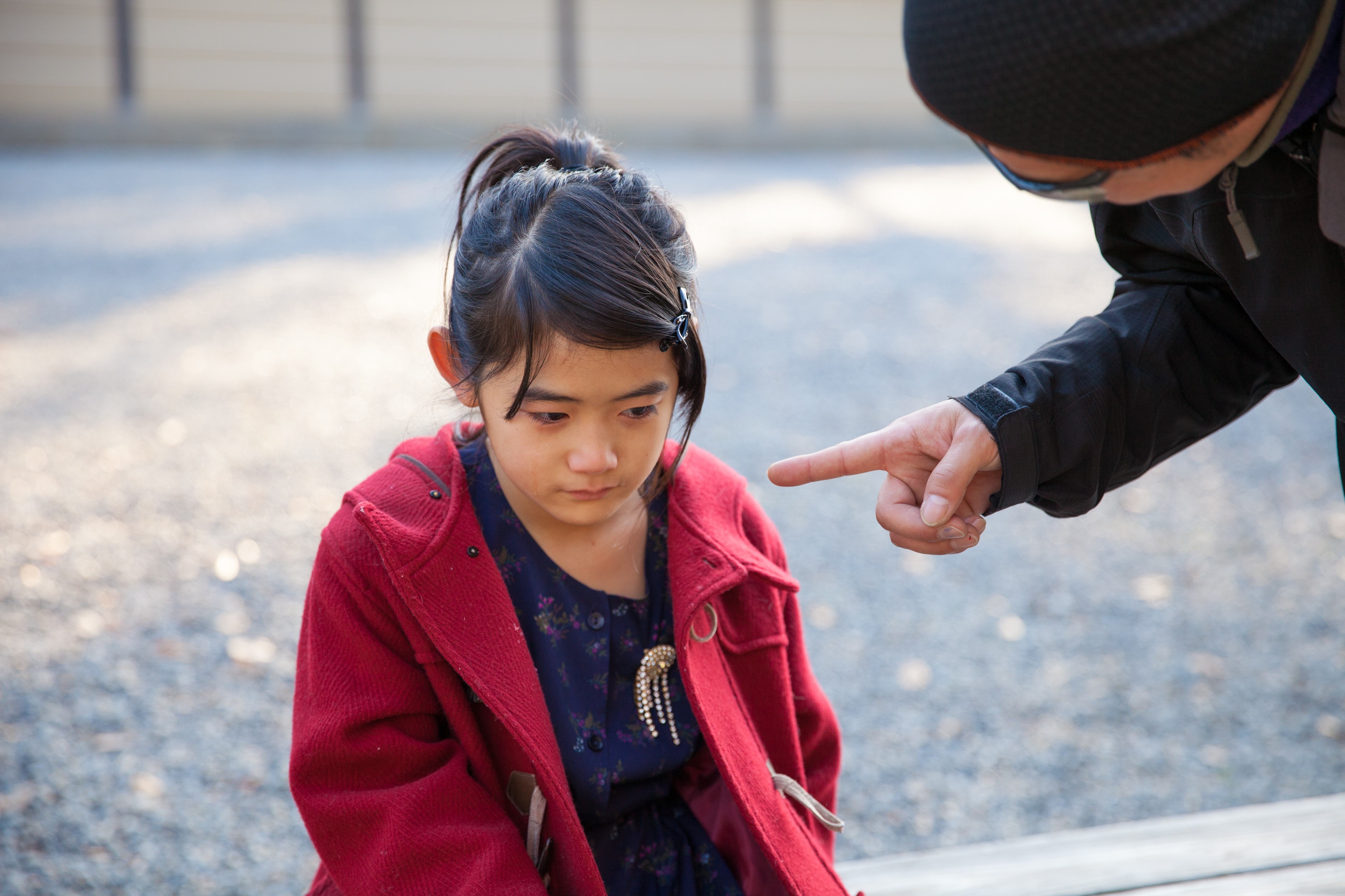 Child in a coat looks down, appearing upset, as adult points at them, suggesting a moment of discipline or guidance in a public space