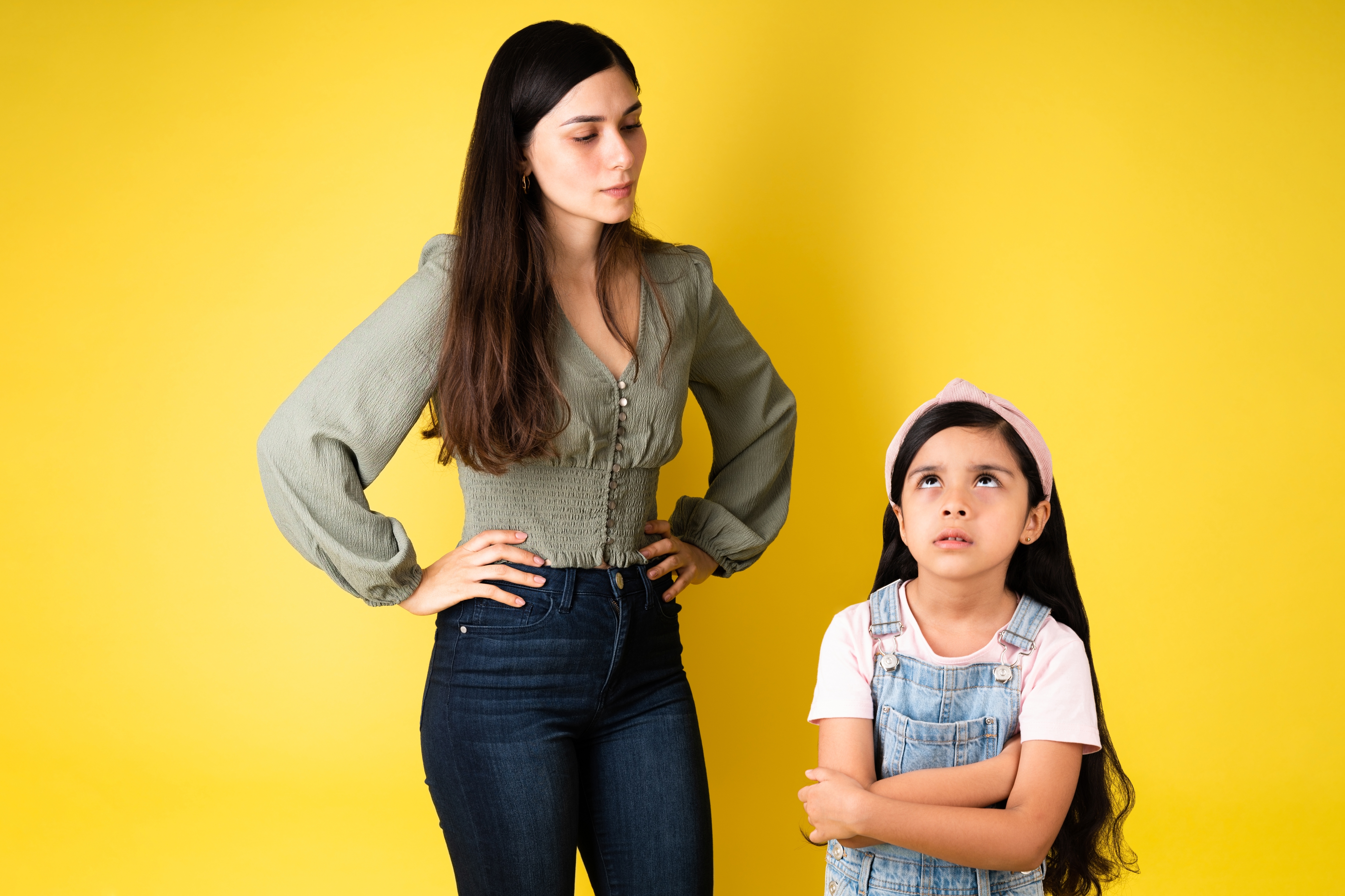 Adult with hands on hips looks at child crossing arms and rolling eyes, both standing against a plain backdrop