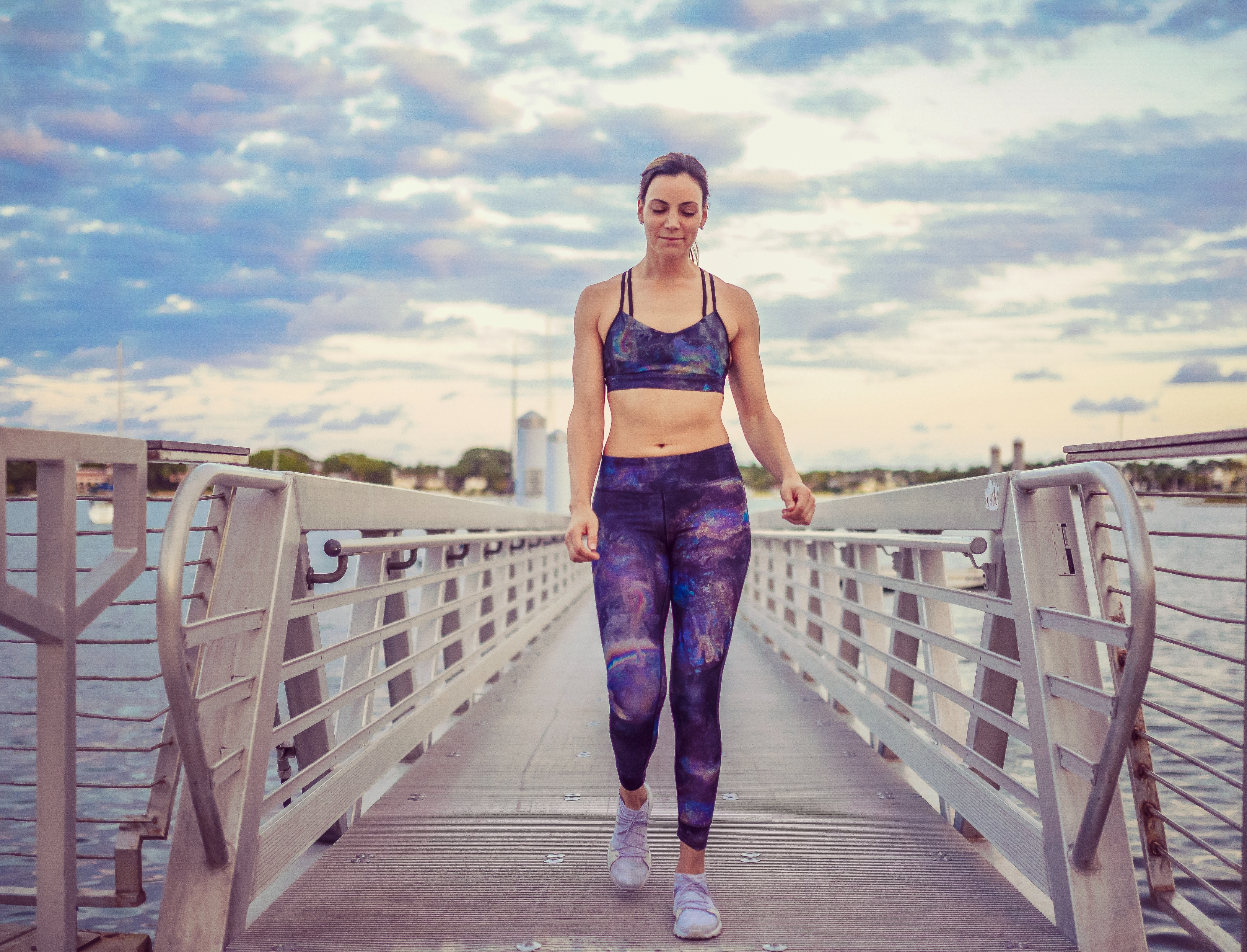Person in fitness attire walks confidently on a bridge with water and sky in the background