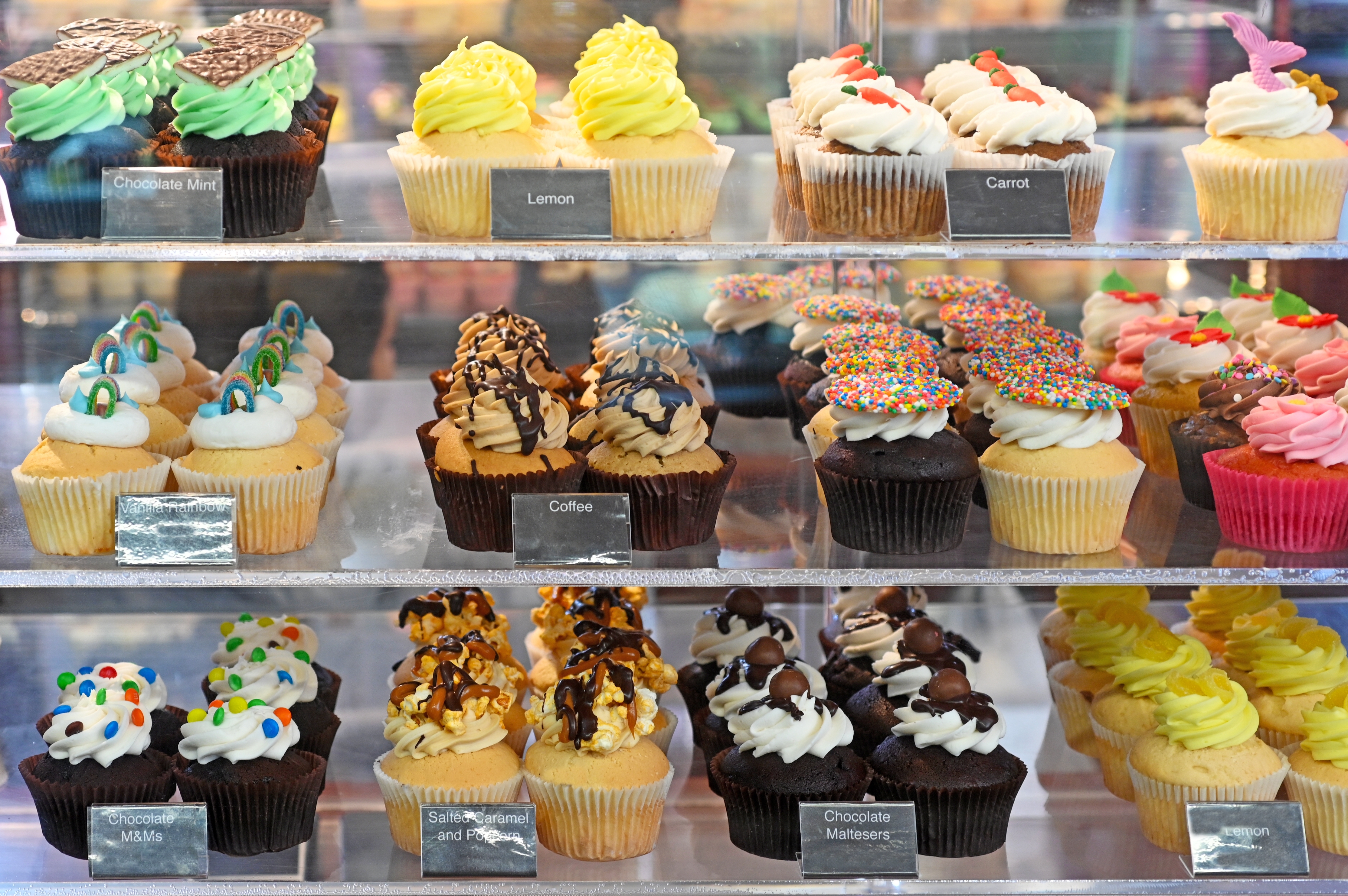 Display of various cupcakes with different toppings and labels, including lemon, coffee, and chocolate varieties, on a bakery shelf