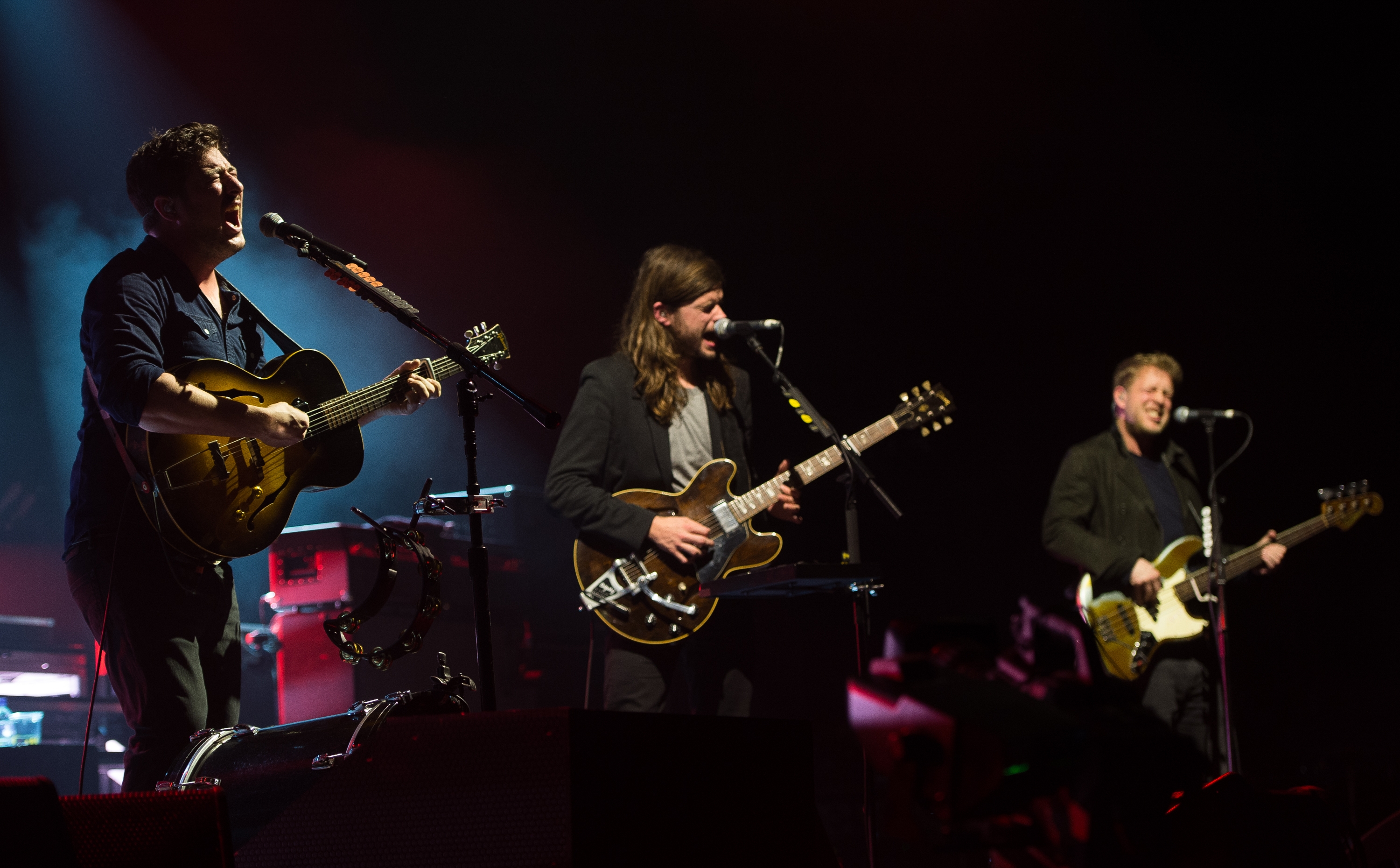 Musicians performing energetically on stage, each playing guitar. The setting is a live concert with visible stage equipment and lighting