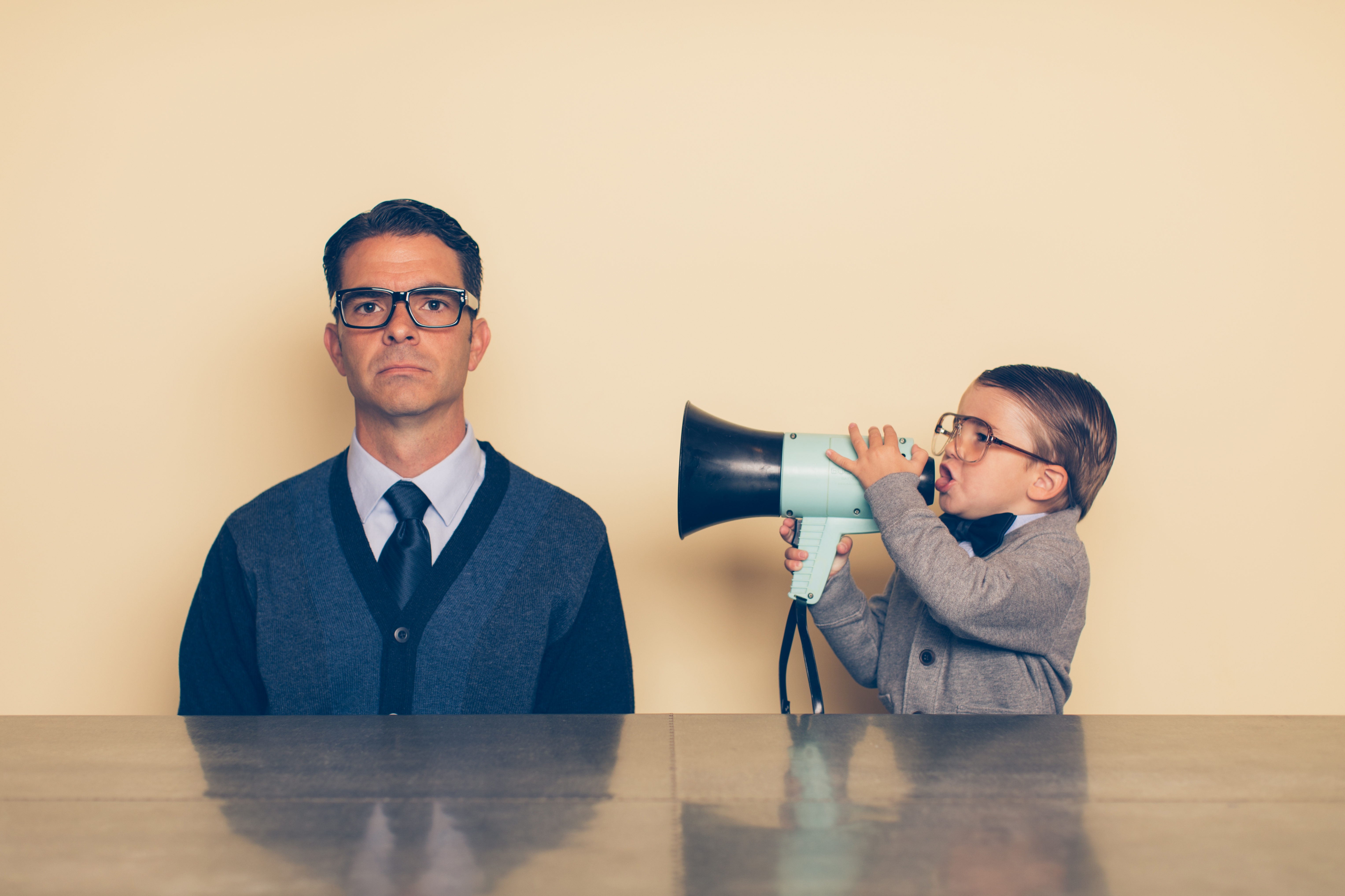 A child playfully uses a megaphone next to a serious-looking man in glasses and formal attire, creating a humorous contrast in a parenting-themed setting