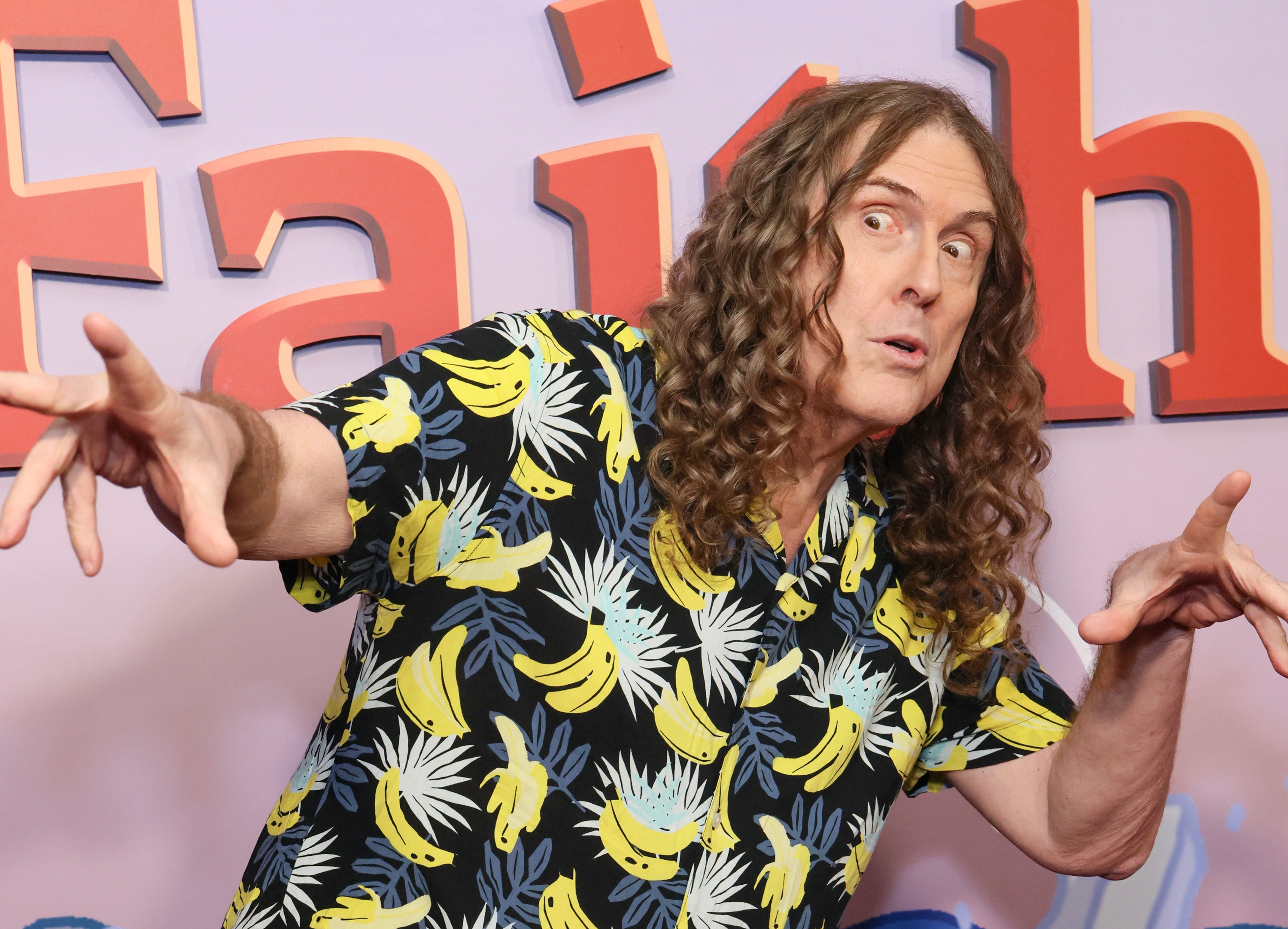 Person in a patterned shirt with bananas and palm leaves poses playfully against a backdrop with the word &quot;Faith.&quot;