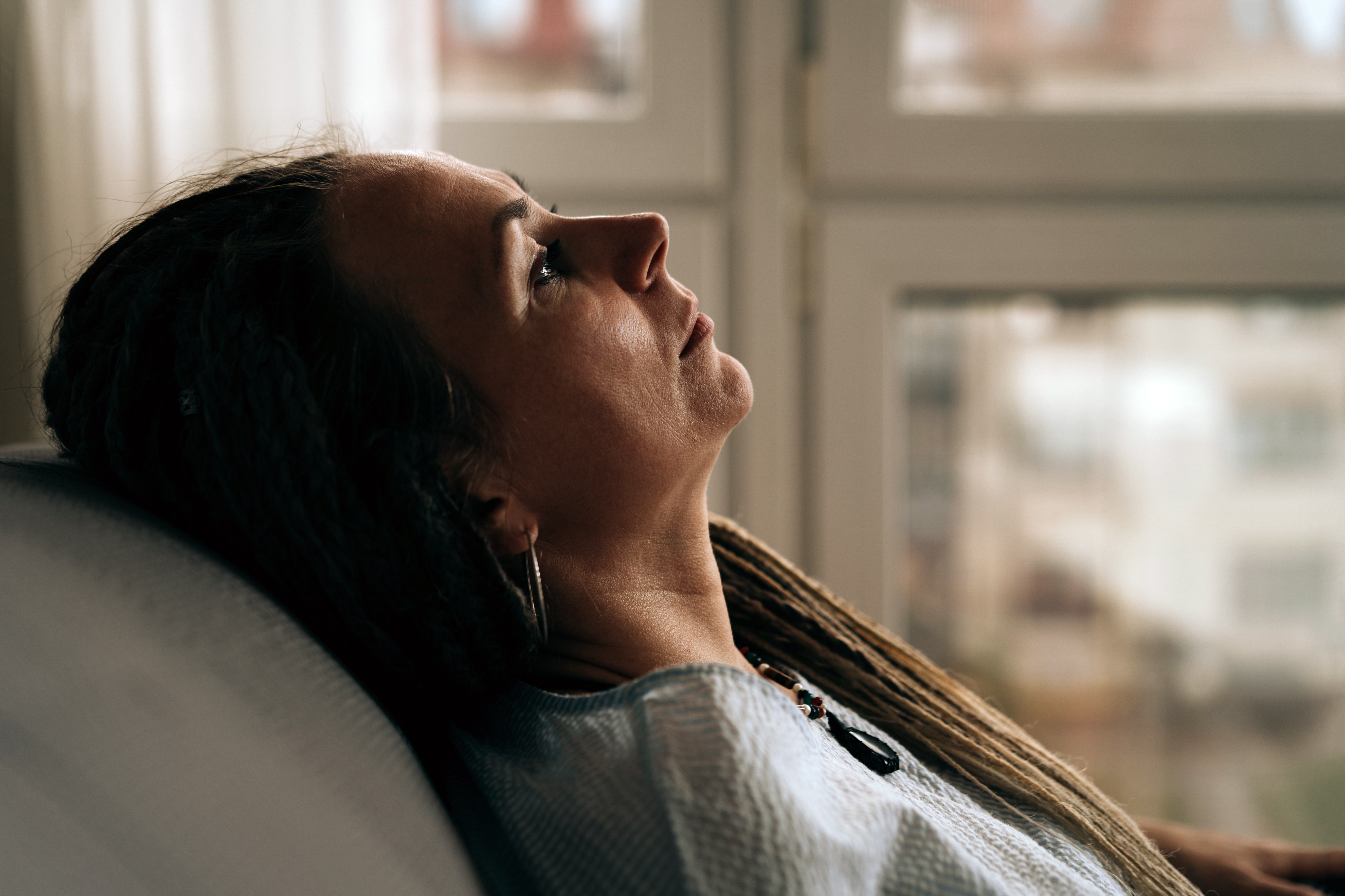 Person with long hair and earrings leans back on a couch, gazing thoughtfully out a window