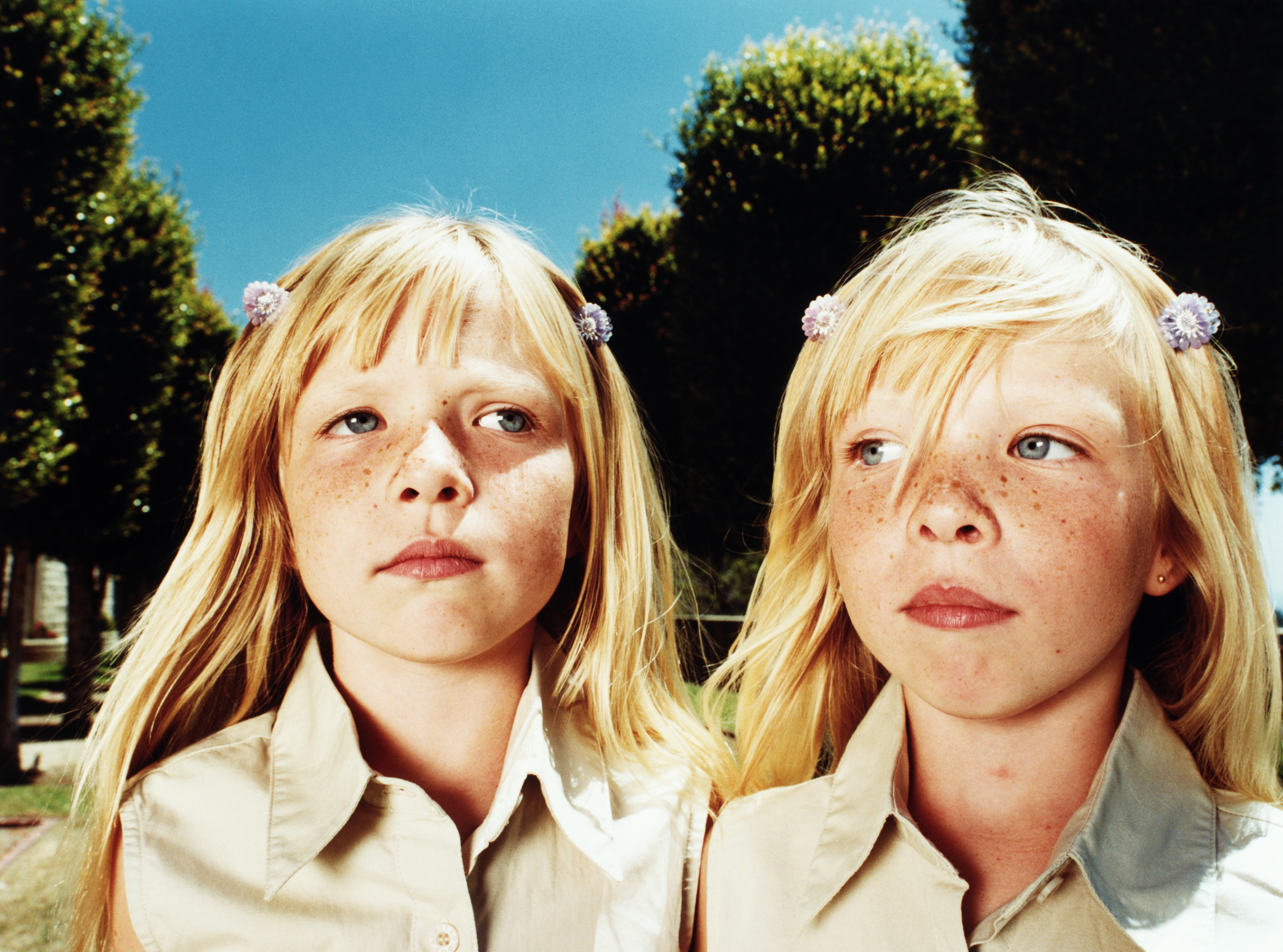 Two young girls with long hair, wearing matching collared shirts, stand outdoors. Trees are visible in the background